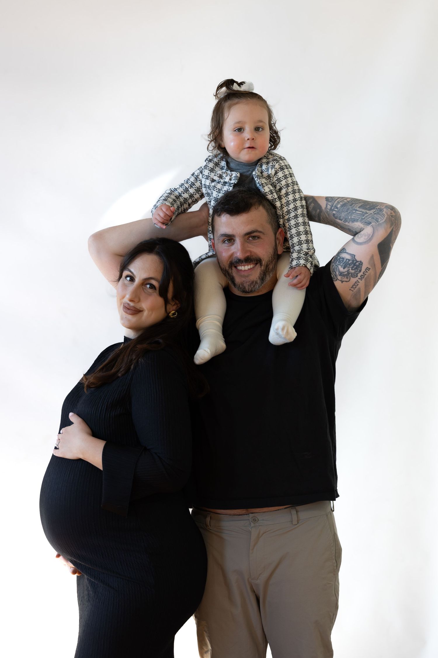 Pregnant woman in black dress poses with tattooed man carrying toddler on shoulders against white background.