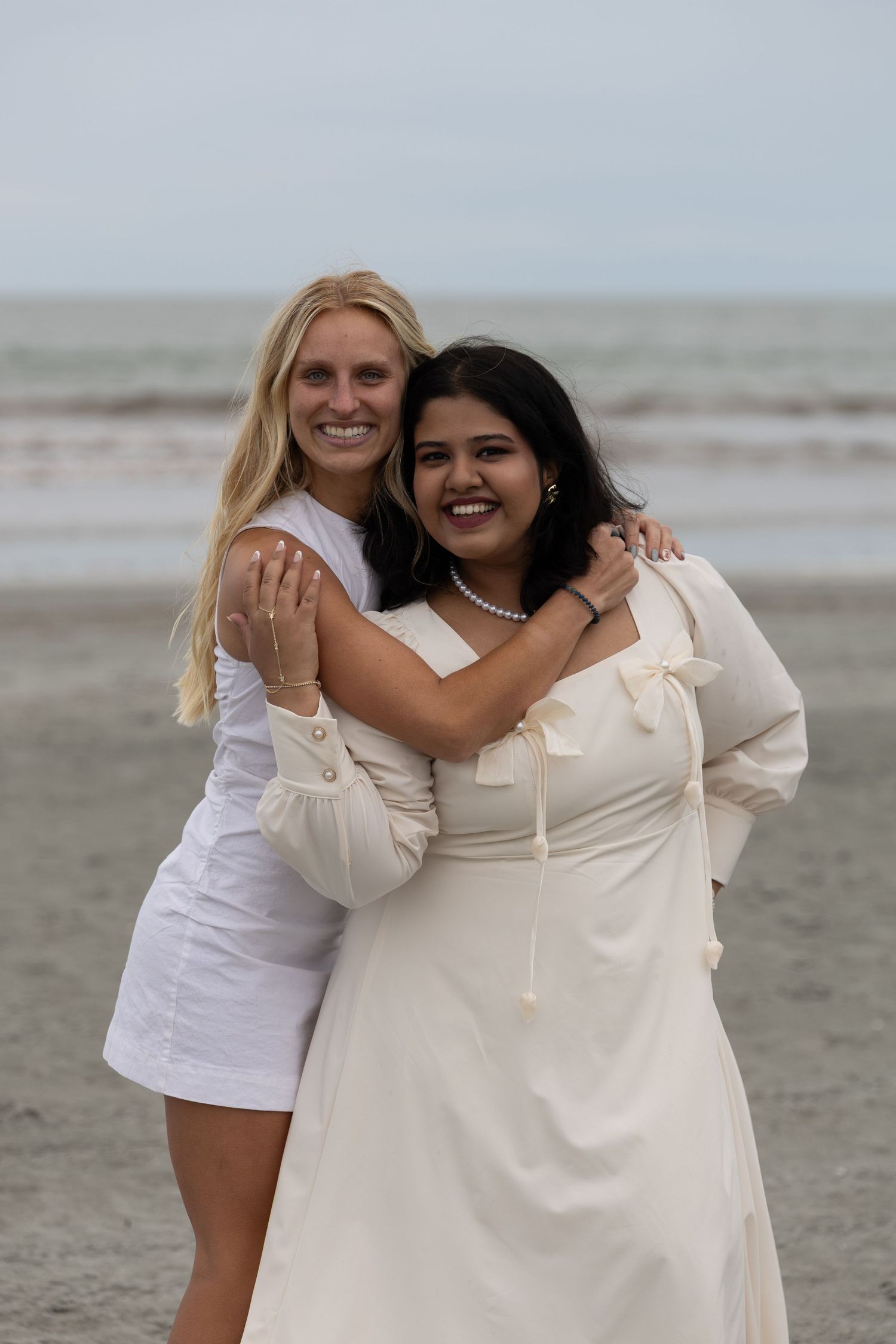 Two women in white dresses smile and embrace on a beach with ocean waves in the background.