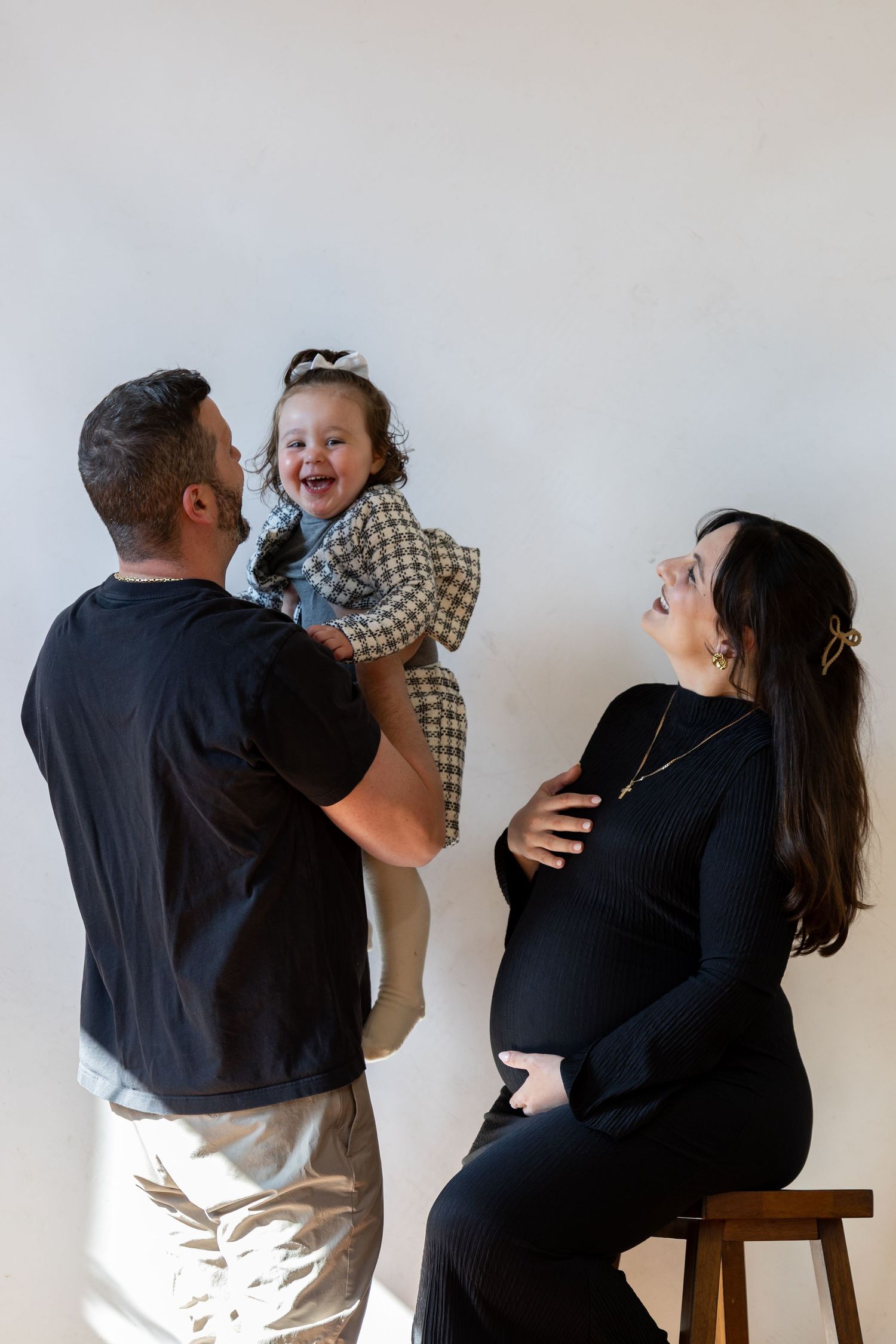 A man lifts a laughing toddler girl while a pregnant woman in black sits smiling on a stool against a white wall.