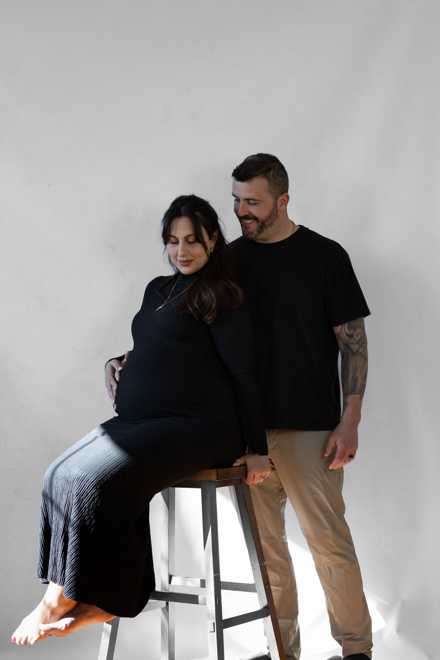 A pregnant woman in black sits on a stool as her smiling partner with tattoos stands beside her in a studio.