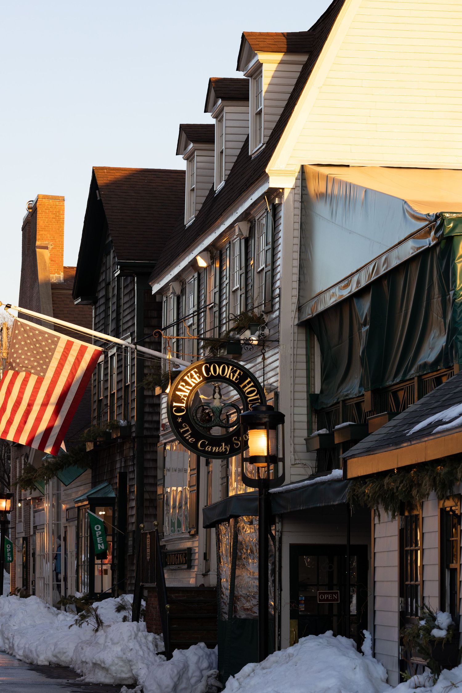 Historic street scene in winter with American flags hanging from buildings and snow lining the sidewalks at sunset.