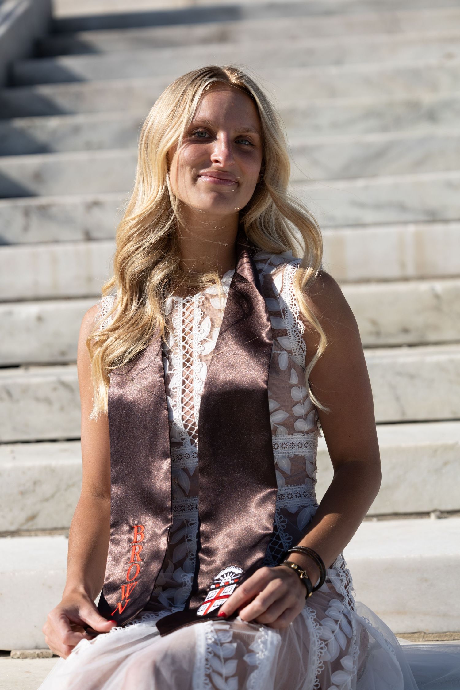 Blonde woman in sleeveless white dress with brown satin sash sits on marble steps, smiling in sunlight.