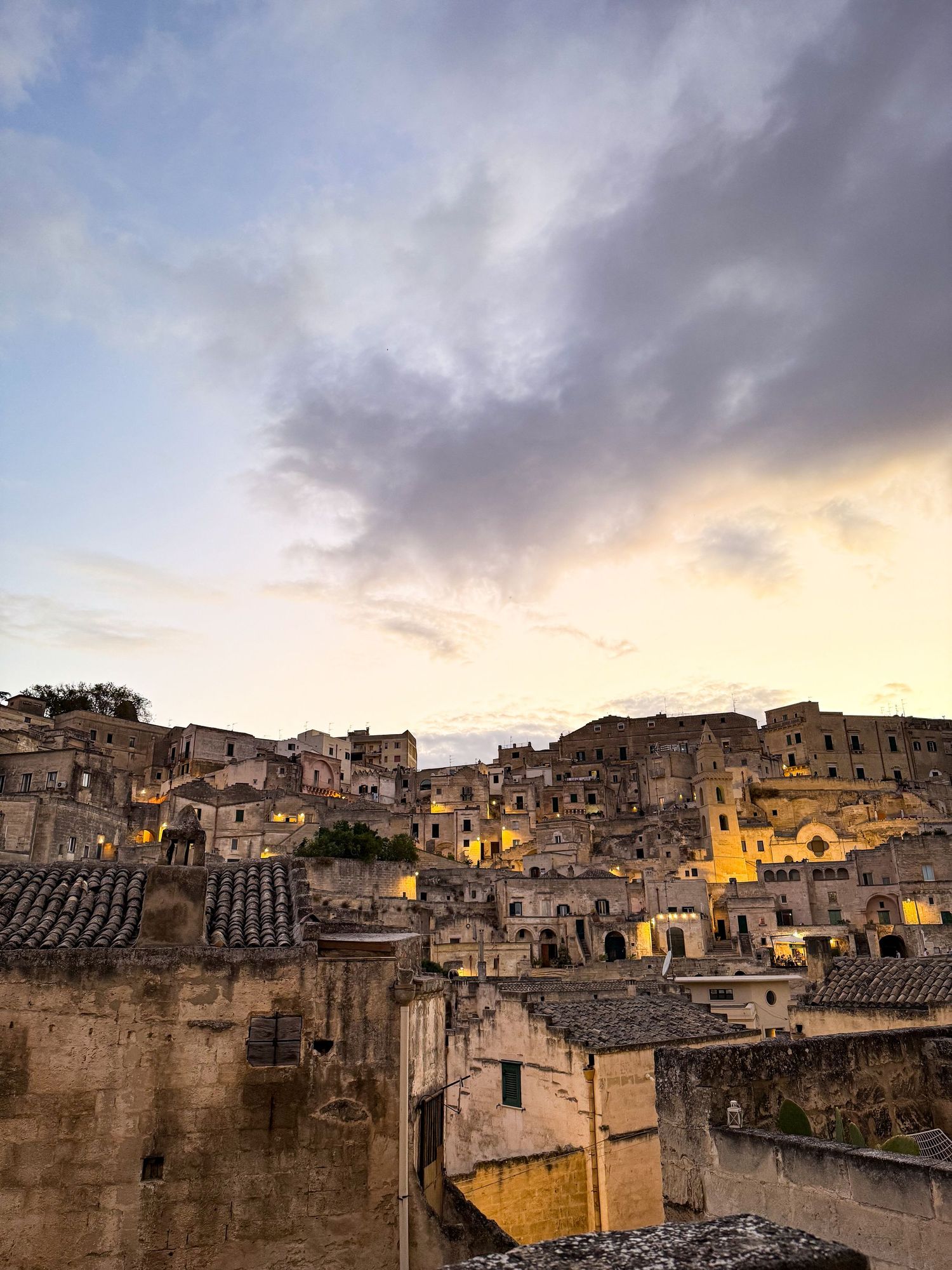 Historic Italian hillside town illuminated at dusk with dramatic cloudy sky overhead.