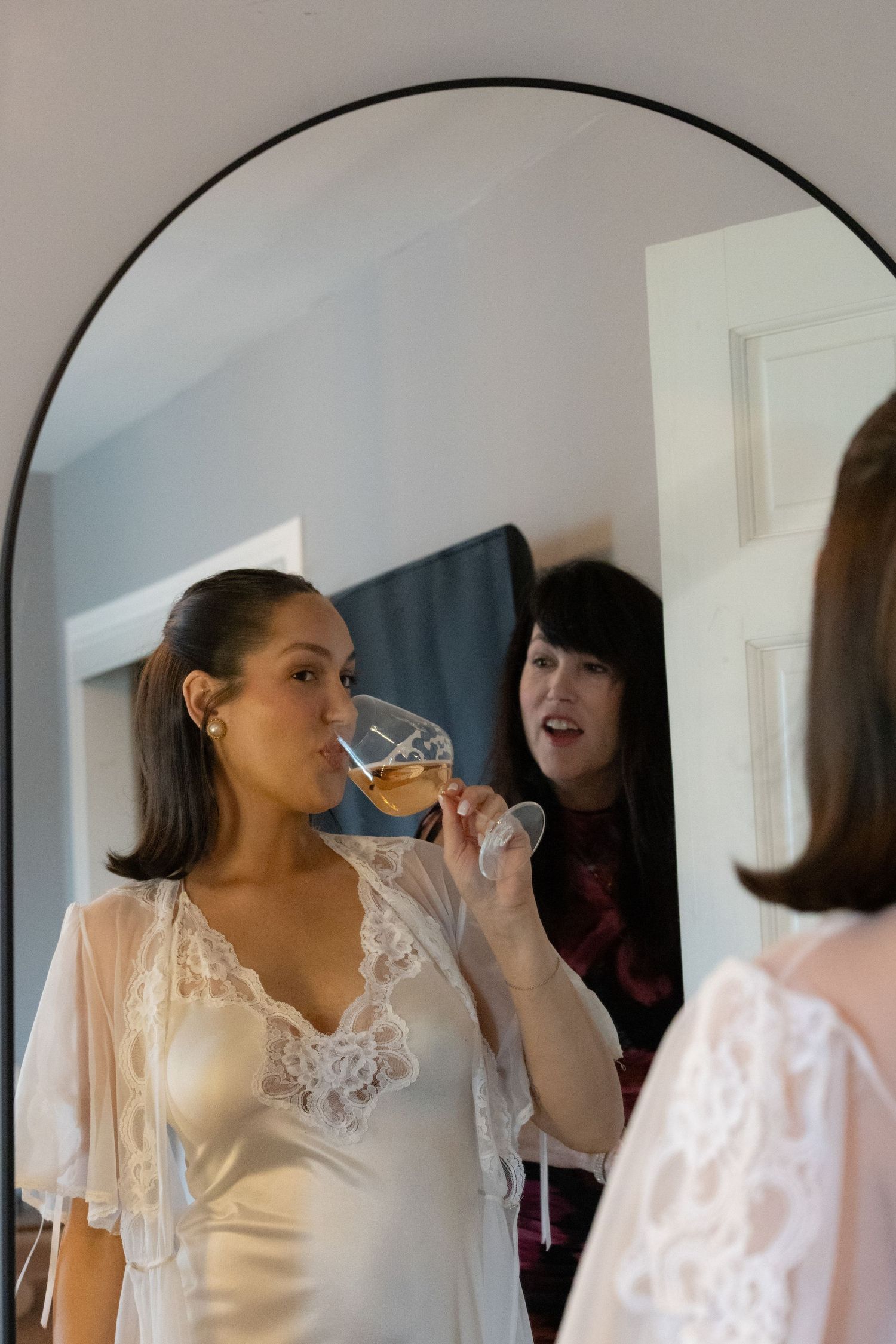 Bride in white lace robe sips champagne while friend watches in arched mirror reflection during wedding preparations.