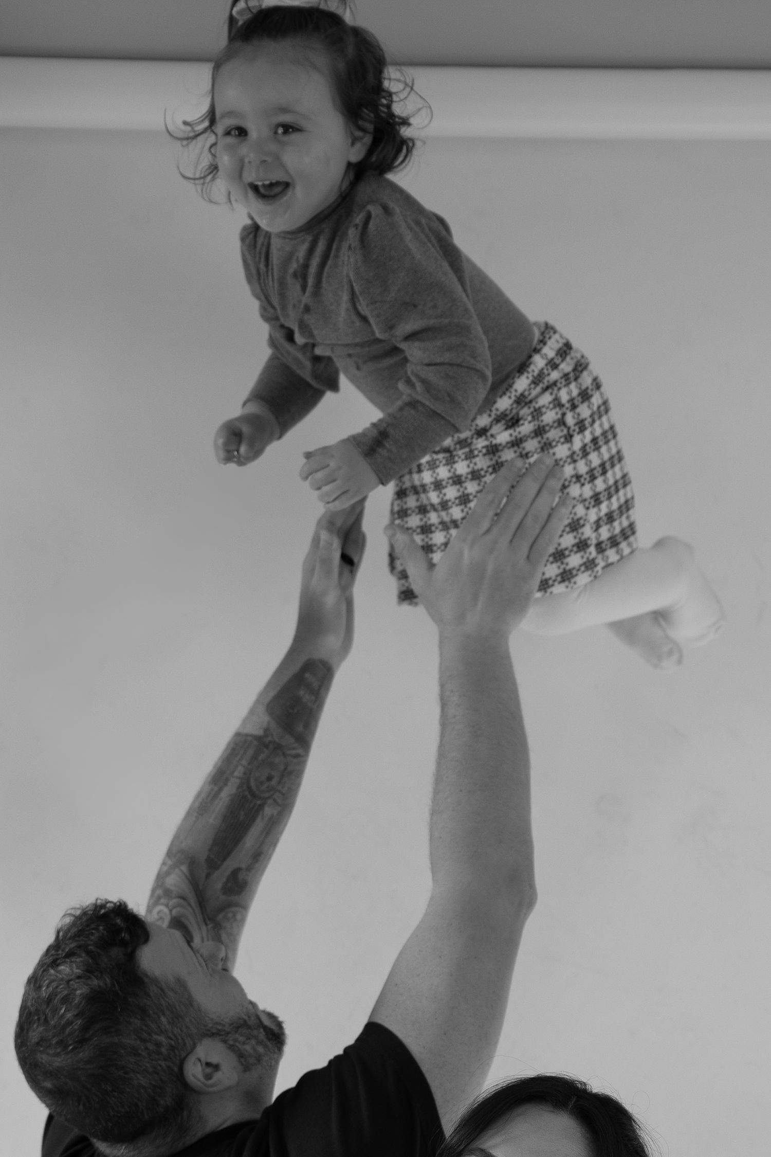 Black and white photo of a tattooed father lifting his laughing toddler daughter high in the air indoors.