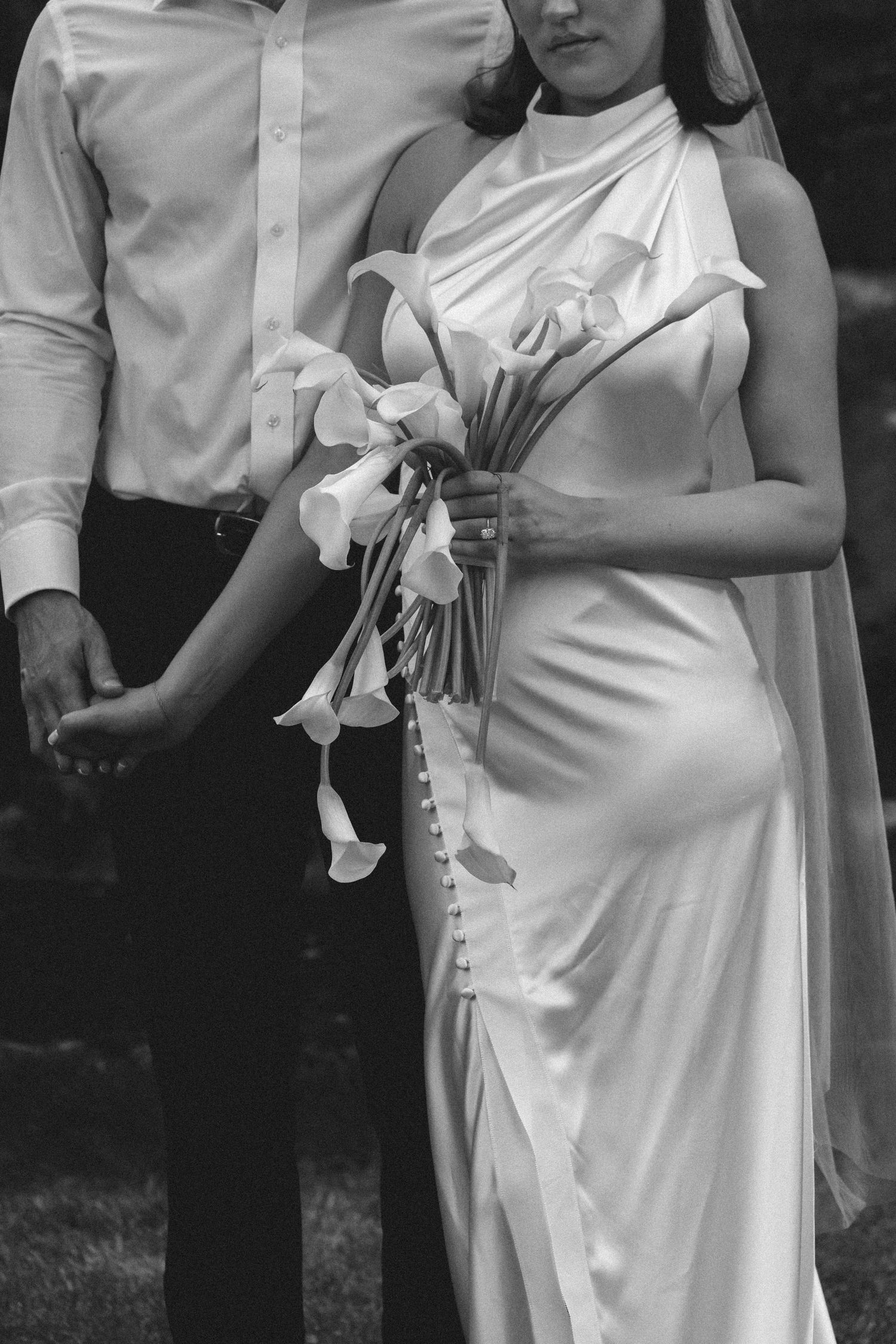 Black and white photo of pregnant bride in silk gown holding calla lily bouquet beside groom.
