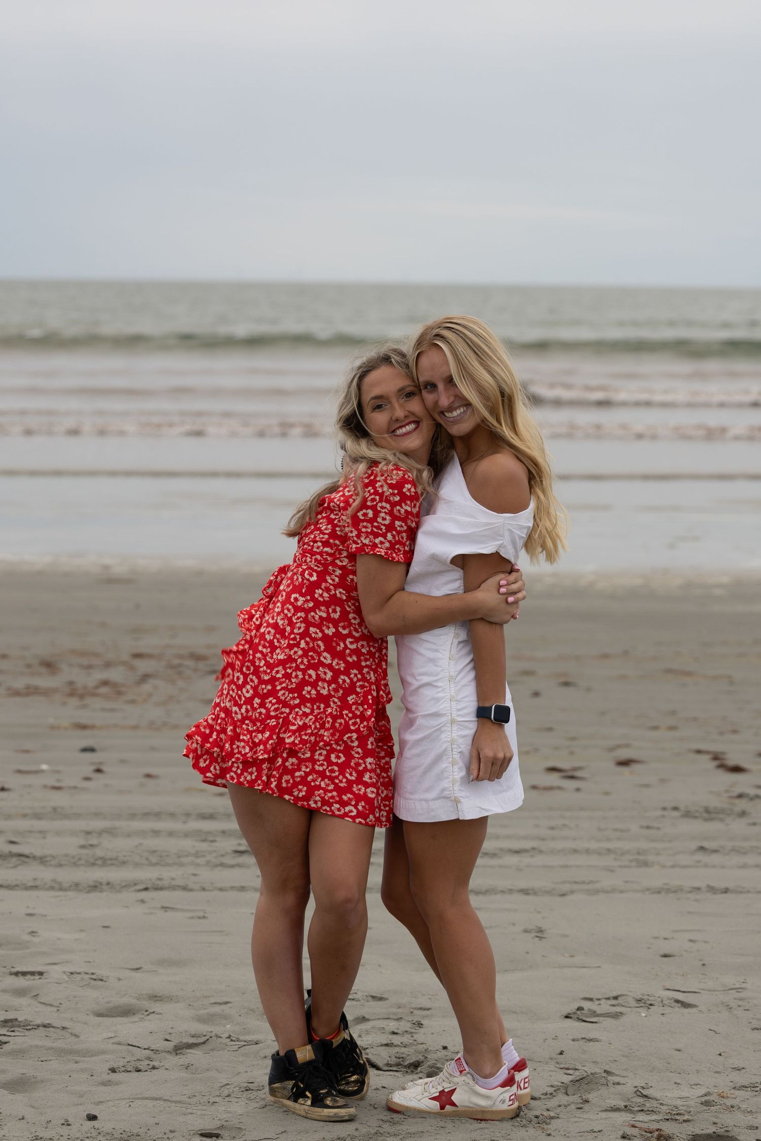 Two blonde women in red and white dresses hugging and smiling on a sandy beach.
