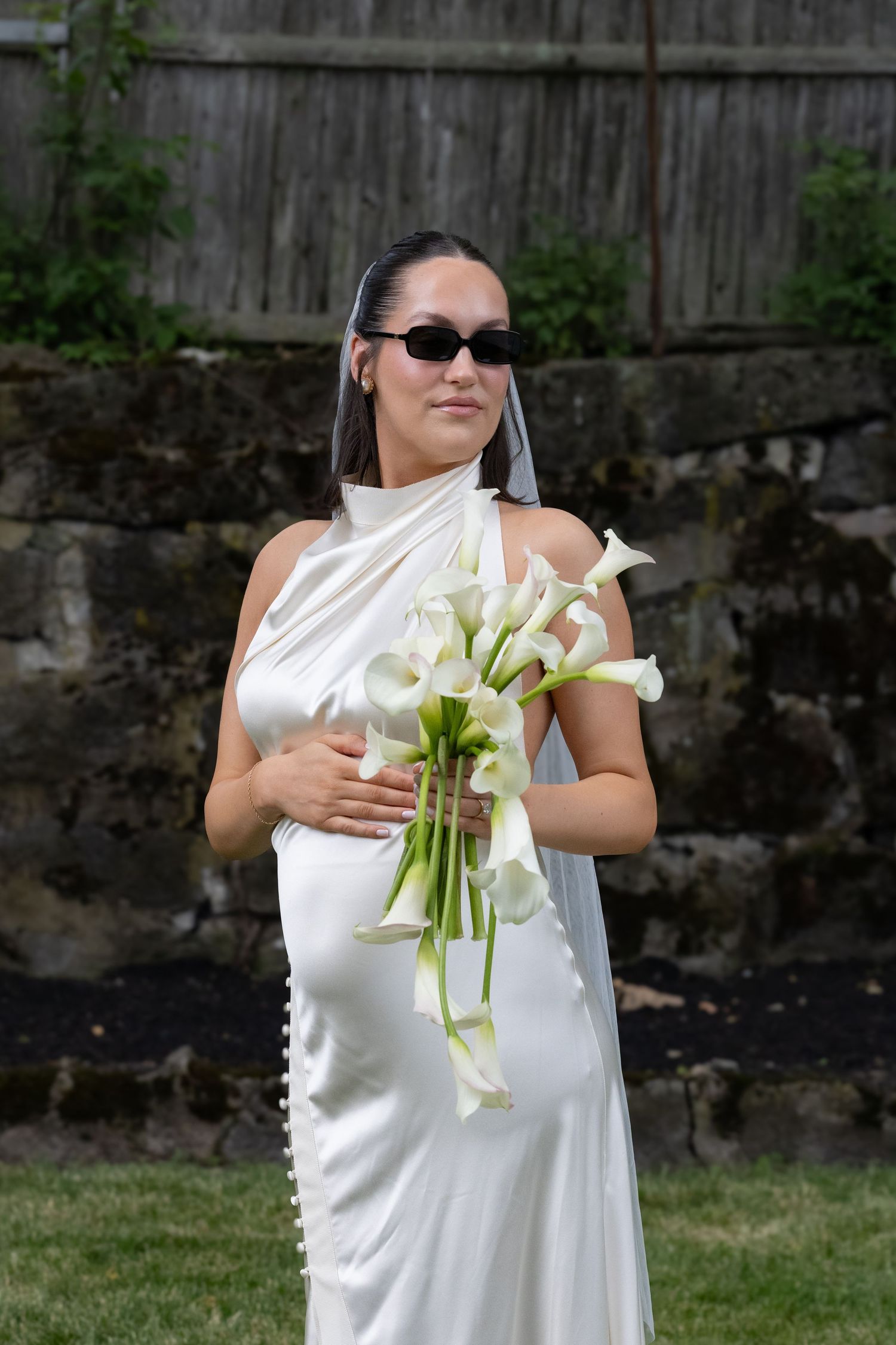 Pregnant bride in white satin halter dress with sunglasses holding calla lily bouquet outdoors.