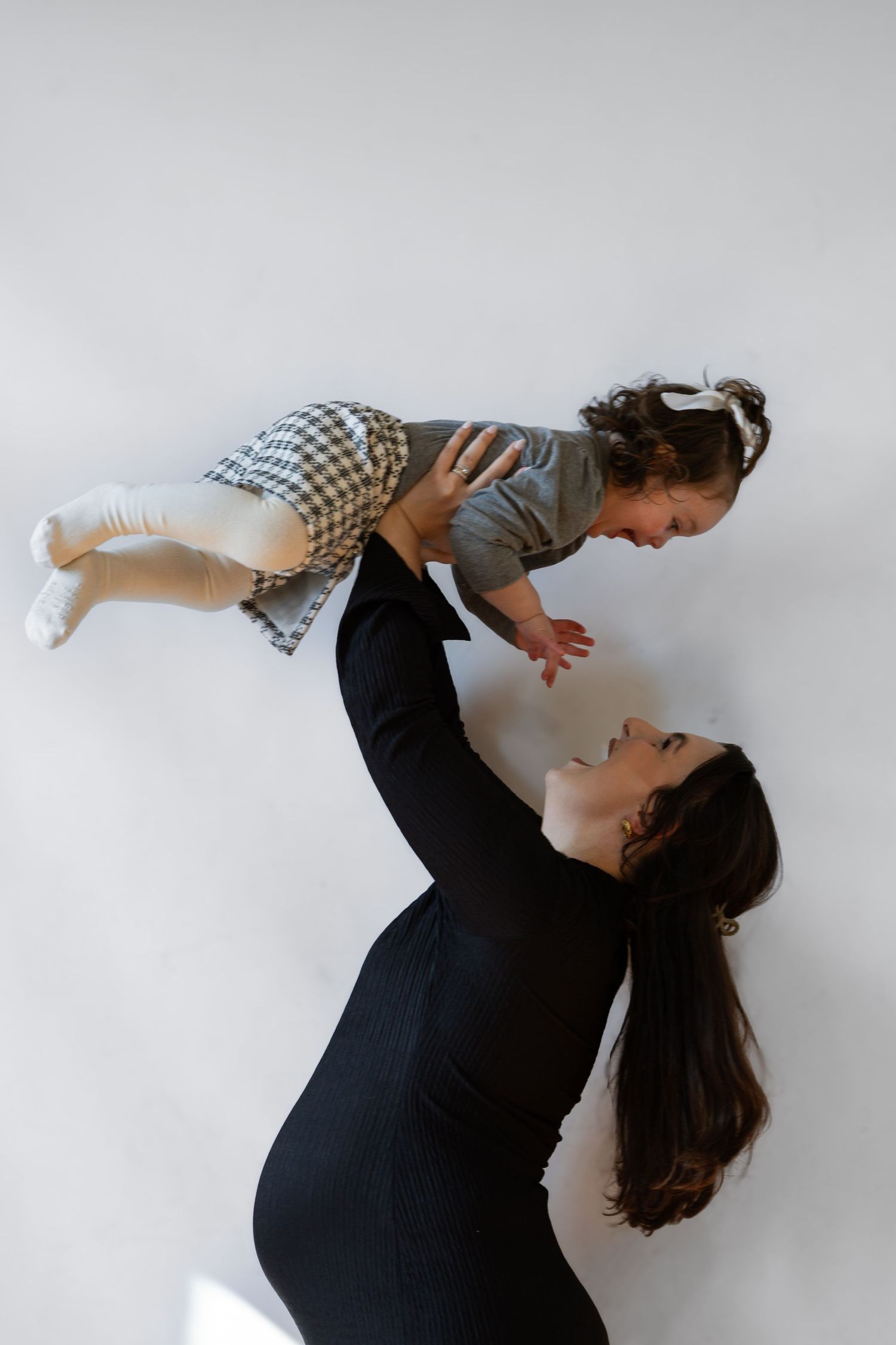 Pregnant woman in black dress lifting smiling toddler overhead against white background.