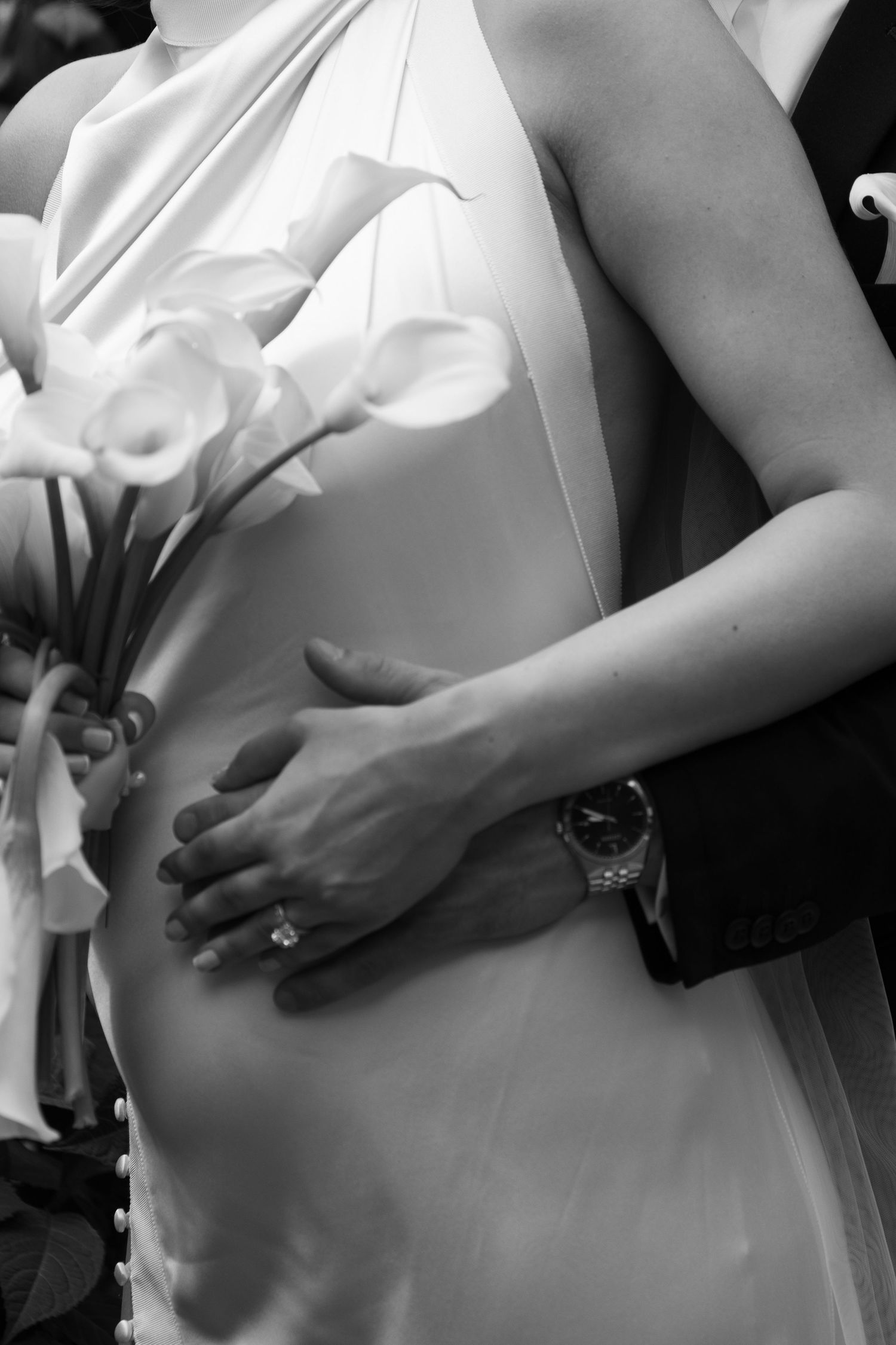 Black and white close-up of bride and groom embracing, showing diamond ring, watch, and calla lily bouquet details.