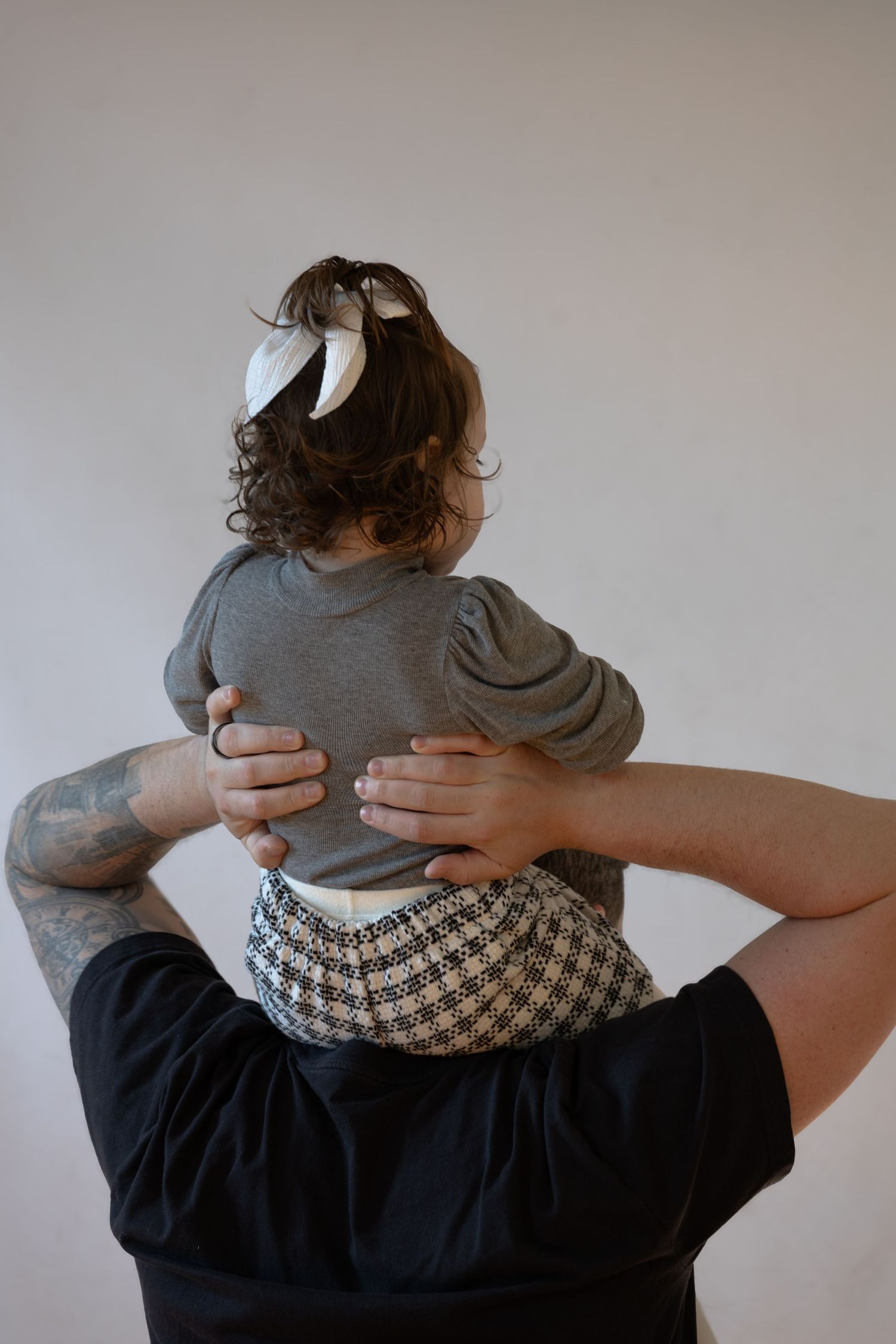 A tattooed adult holds a young child with curly hair and a white bow on their shoulders, both facing away.