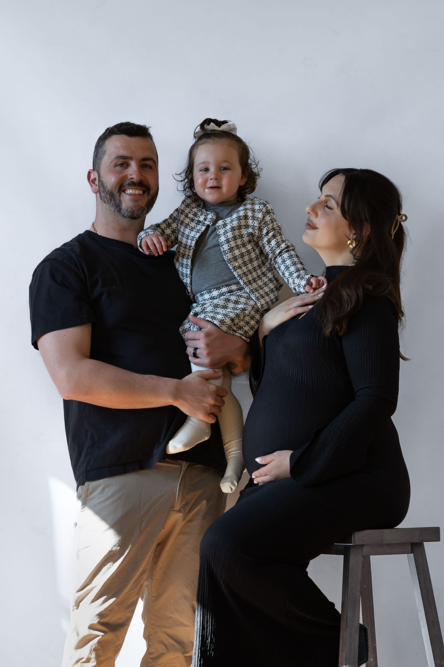 Smiling family maternity photo with dad holding toddler girl and pregnant mom in black dress against white wall.