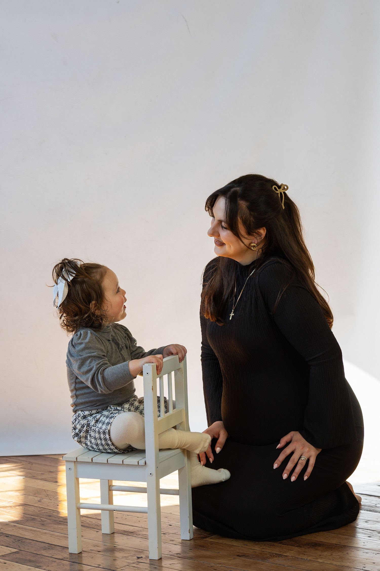 Pregnant mother in black dress kneels on wooden floor interacting with young toddler sitting on tiny white chair.