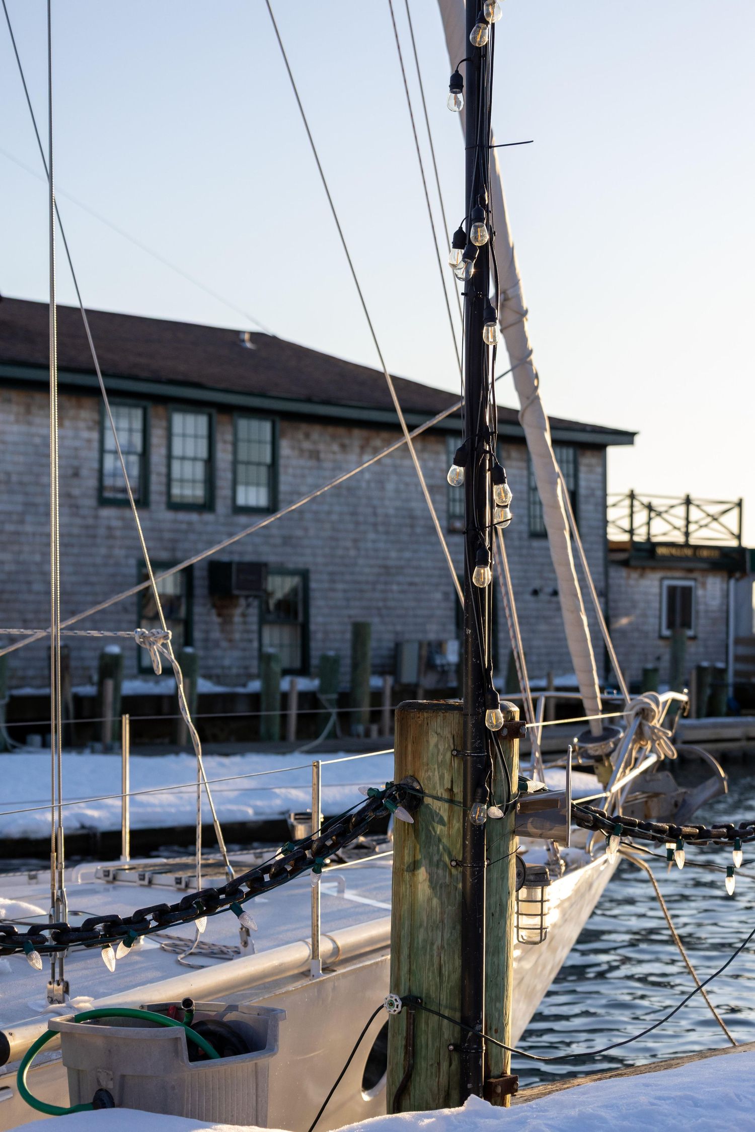 Sailboat masts and rigging line the dock at a marina with wooden pilings and buildings in the background.