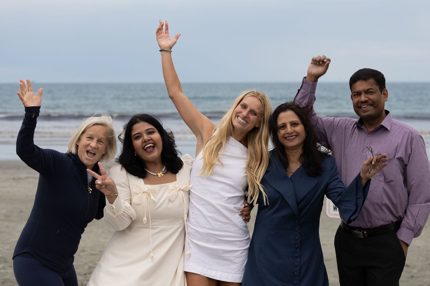 Five diverse friends celebrating on a beach, smiling and raising their arms in joy on a cloudy day.