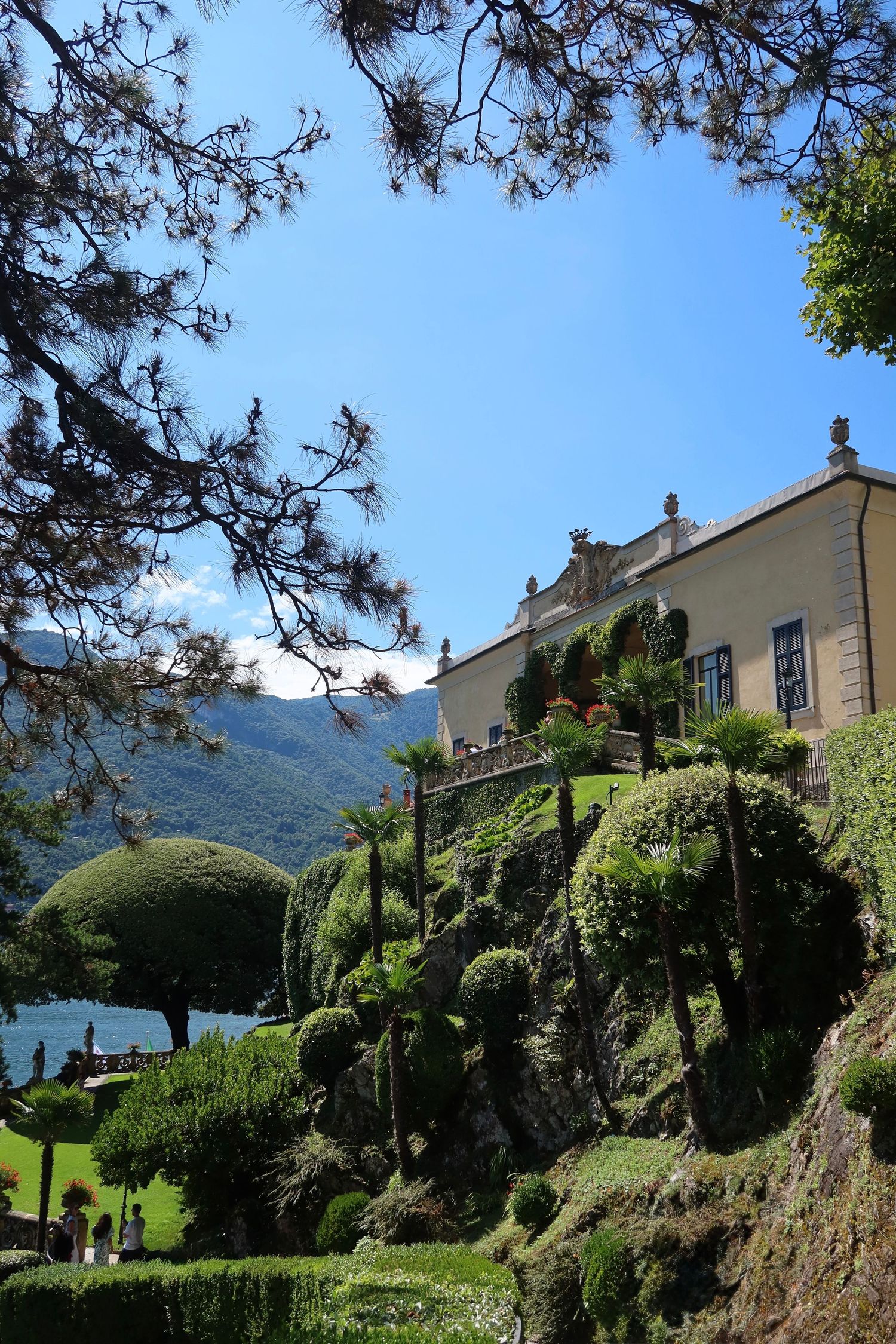 Manicured topiary garden with sculptured bushes and villa buildings overlooking a lake.