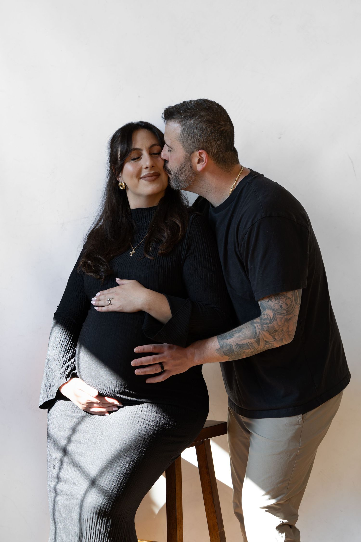 Pregnant woman seated on stool as tattooed husband kisses her cheek, both cradling baby bump, maternity photo.