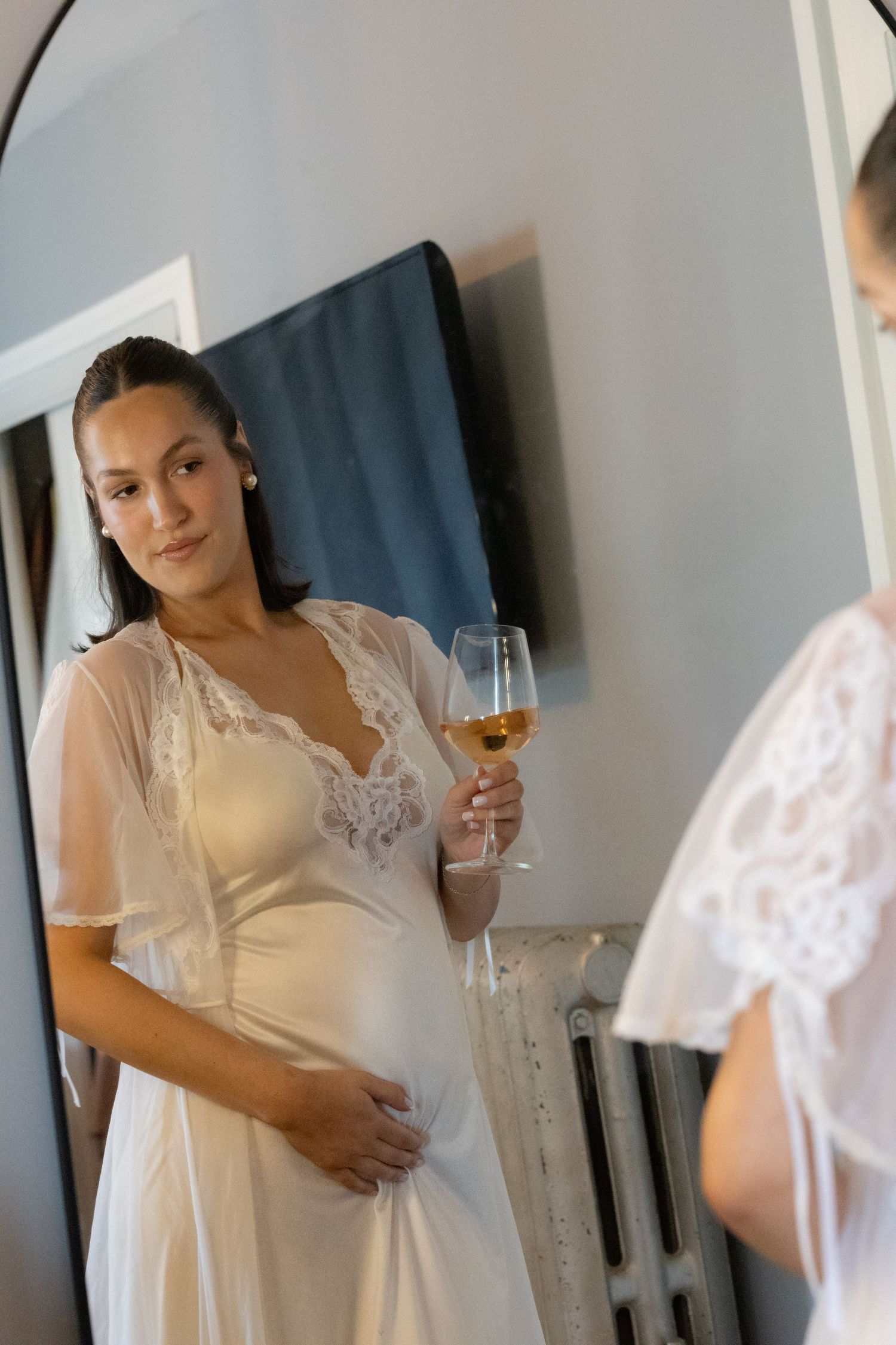 Elegant bride in white lace robe holds champagne glass while admiring her reflection in an arched full-length mirror.