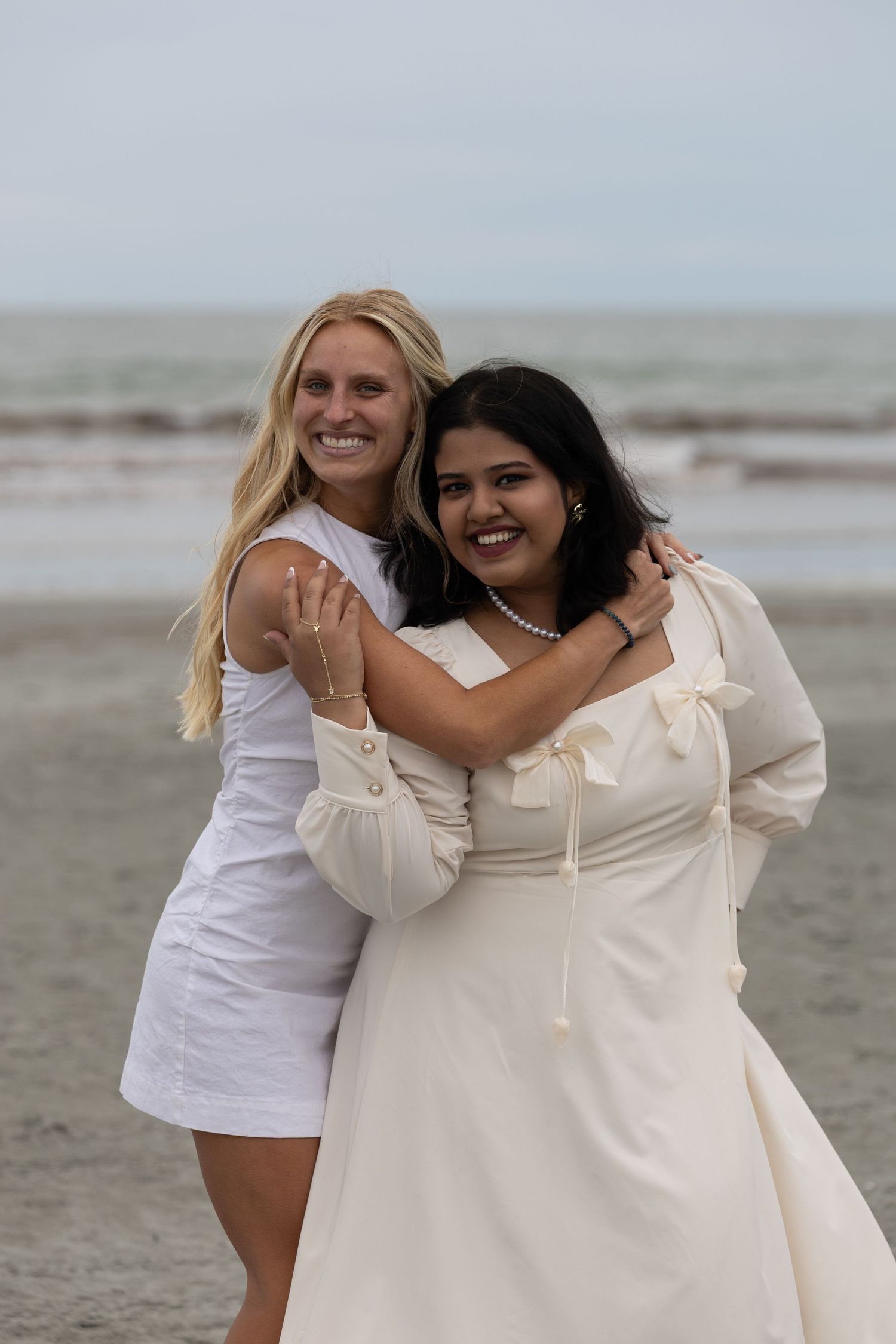 Two women in white dresses hugging and smiling on a beach with ocean in background.