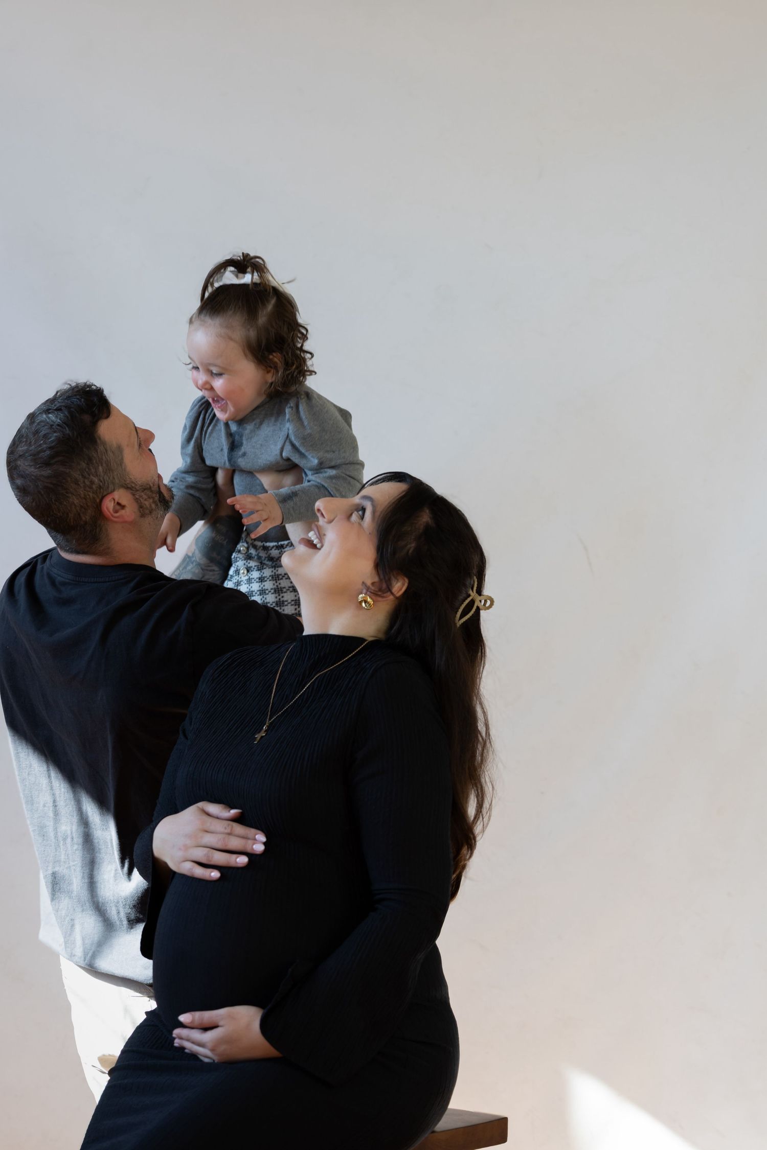 A happy pregnant woman in black sits with her partner holding their laughing toddler daughter.
