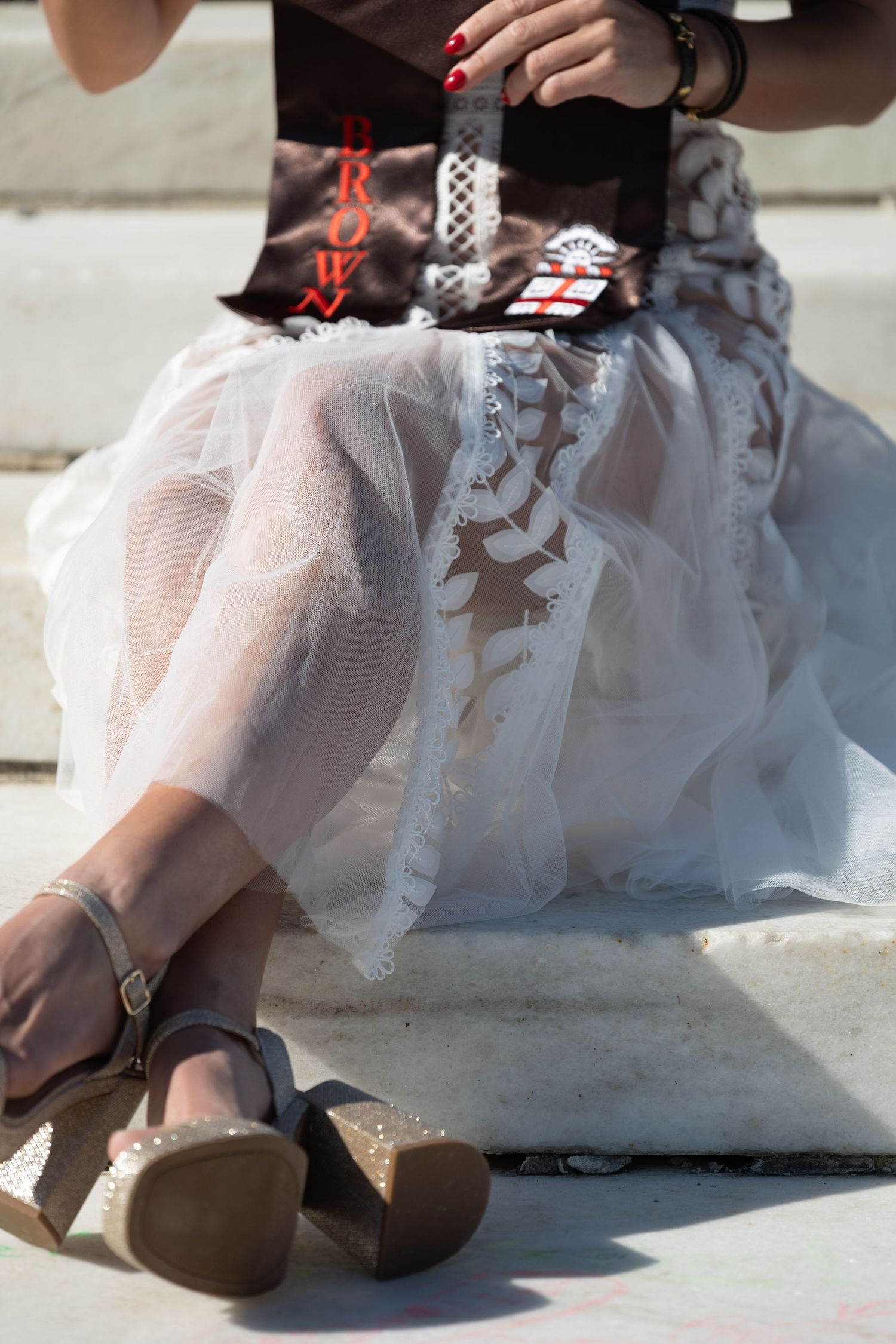 Woman in white tulle skirt and wedge sandals sitting on steps, holding a brown bag with red nails.