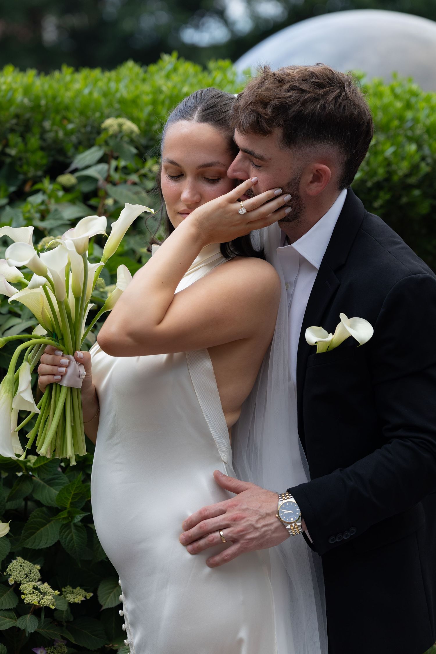A bride in a white gown holding calla lilies embraces a groom in a black suit with a floral boutonniere outdoors.