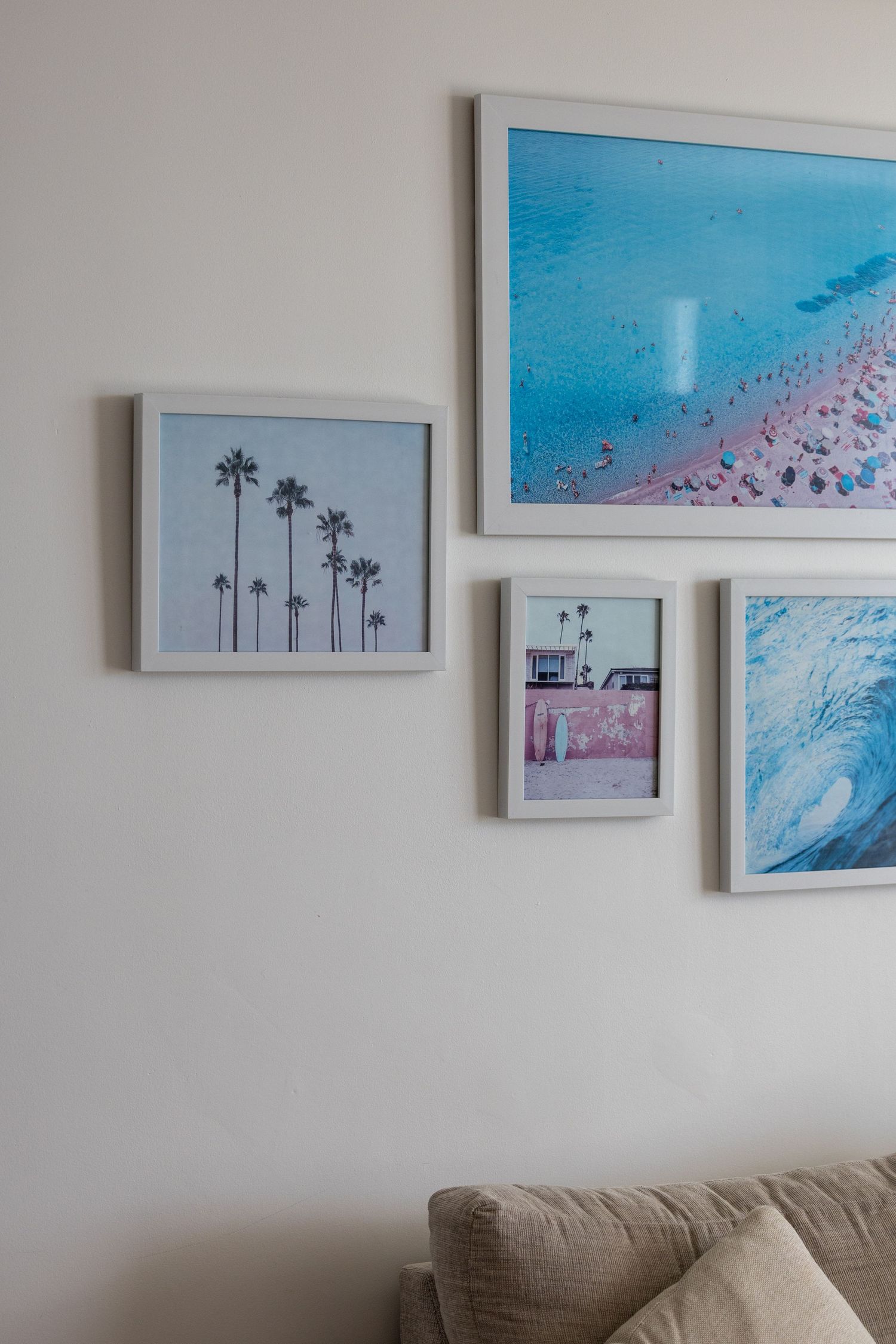Four framed coastal art prints on a white wall above a beige sofa, featuring palm trees, beach aerial, surf shop, and ocean wave.