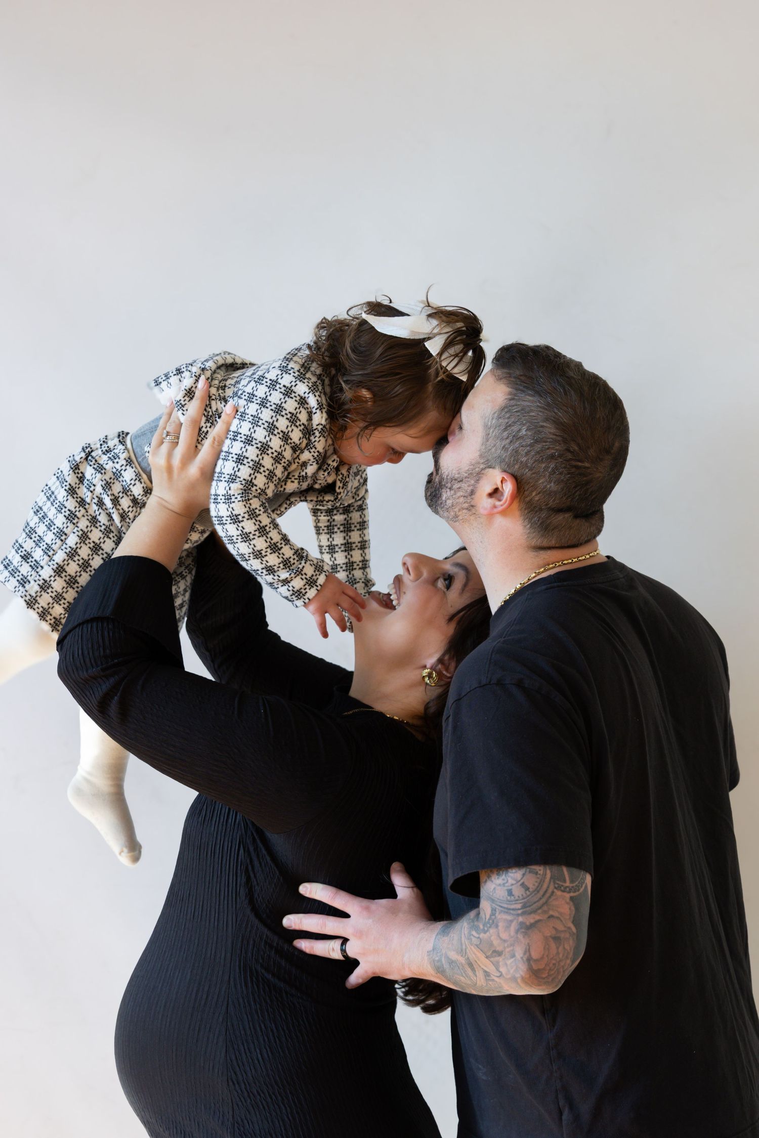 Couple lifting toddler girl in houndstooth dress, pregnant mother in black dress, father with tattoos, family maternity photo.