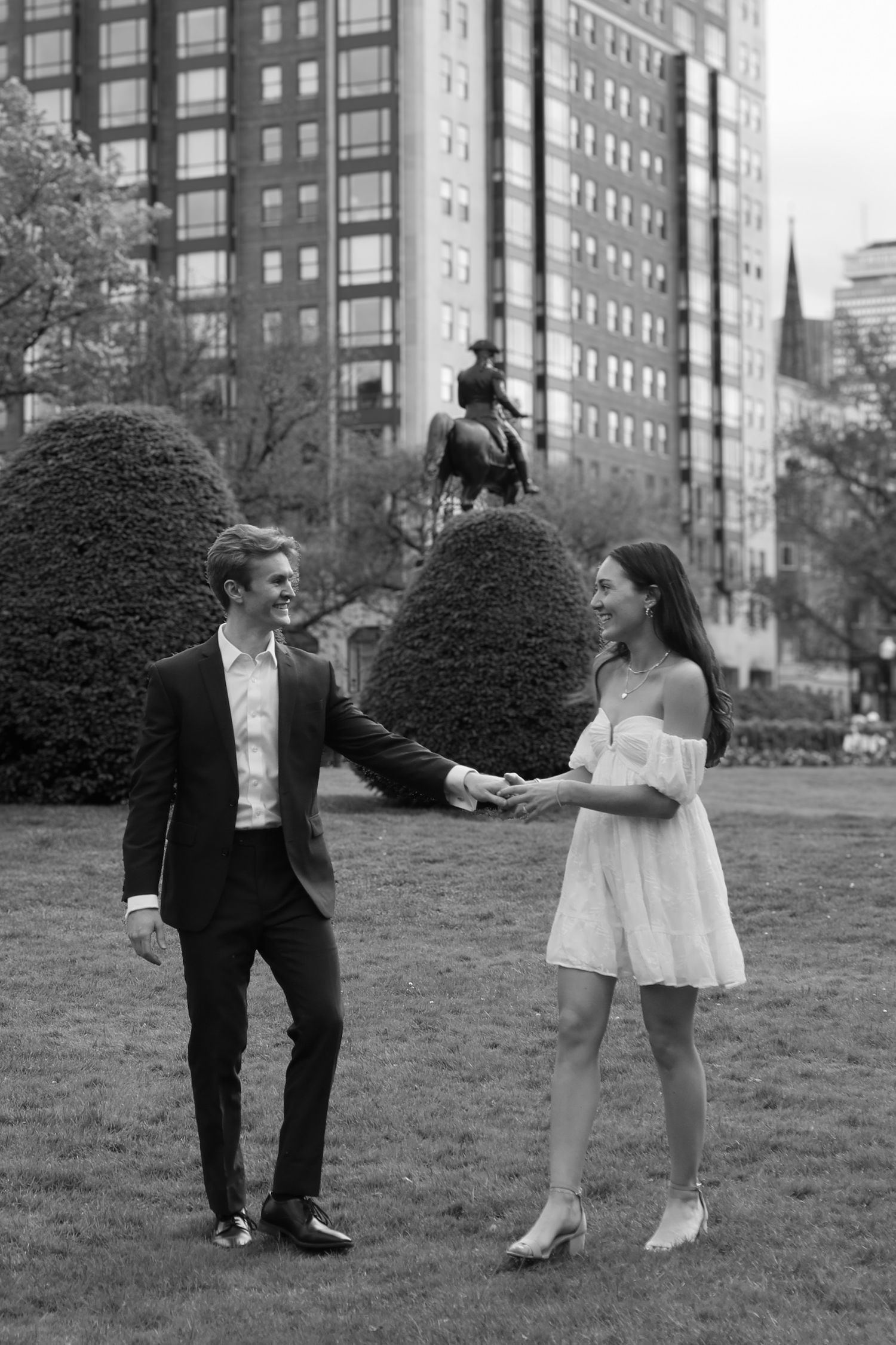A couple holds hands while standing on grass with city architecture in the background.