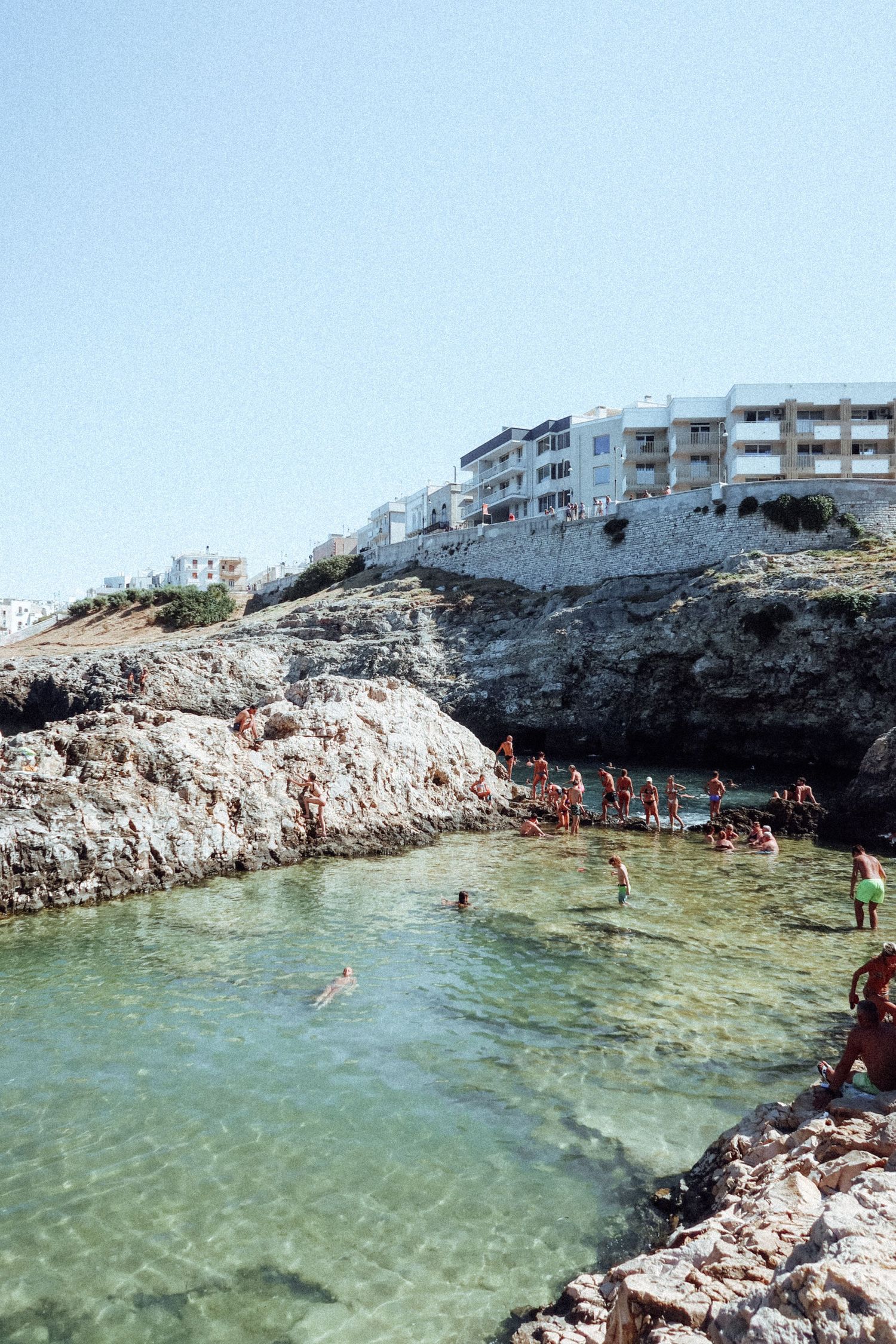 People enjoy swimming and cliff jumping at a natural rocky cove with crystal clear turquoise waters near coastal buildings.