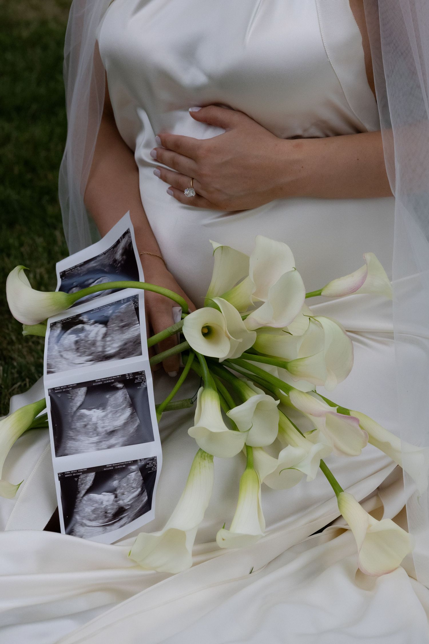 Pregnant bride in white gown with calla lily bouquet and ultrasound photos displayed on wedding dress.