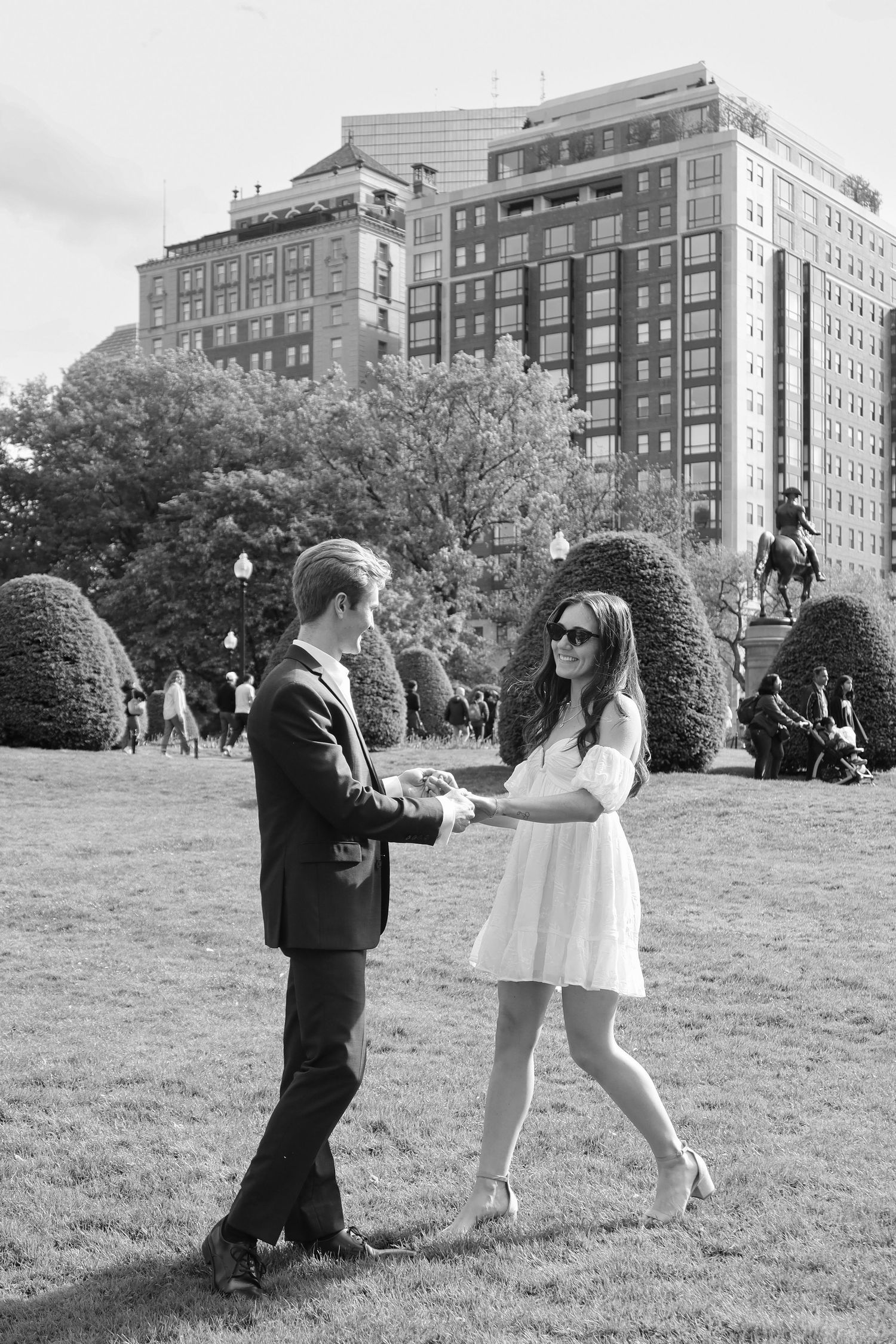 A couple dances together on a lawn surrounded by high-rise buildings in an urban park.
