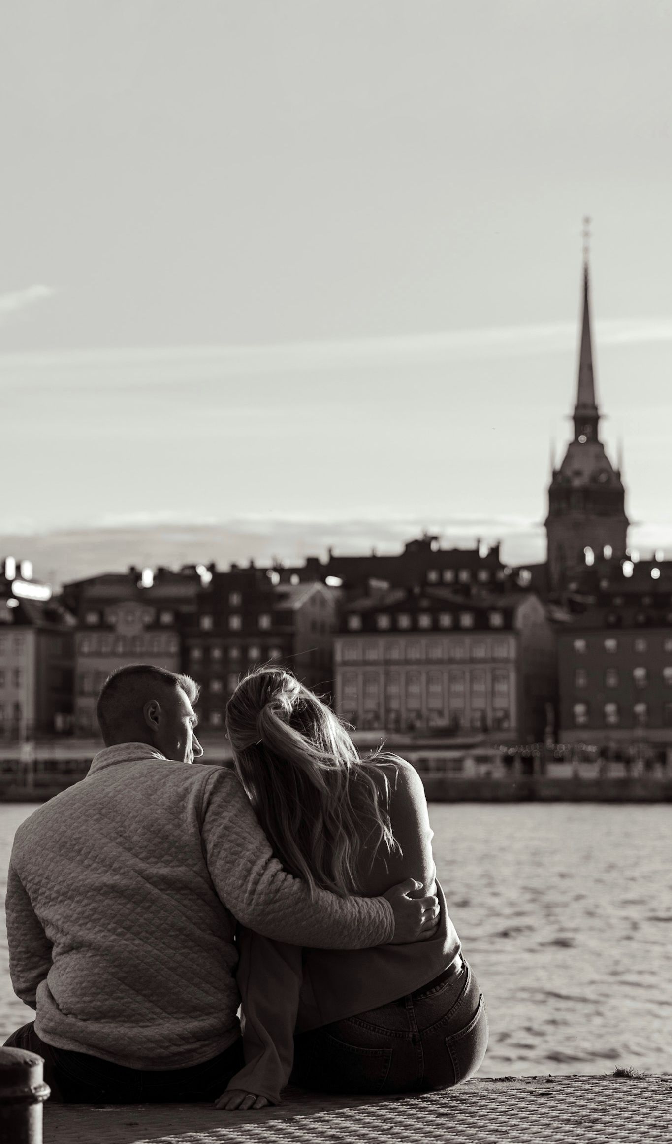 A romantic couple sits together overlooking a waterfront cityscape with church spires in Stockholm, Sweden.