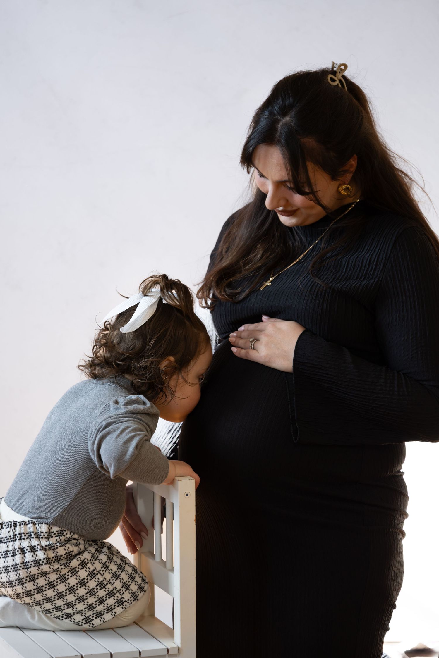 Pregnant woman in black dress with toddler kissing her baby bump during a maternity photo session.