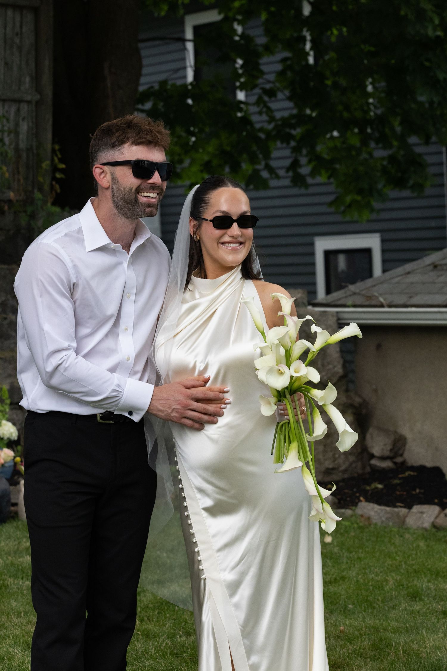 Smiling bride and groom wearing sunglasses pose outdoors, bride holding white calla lily bouquet in elegant satin wedding gown.