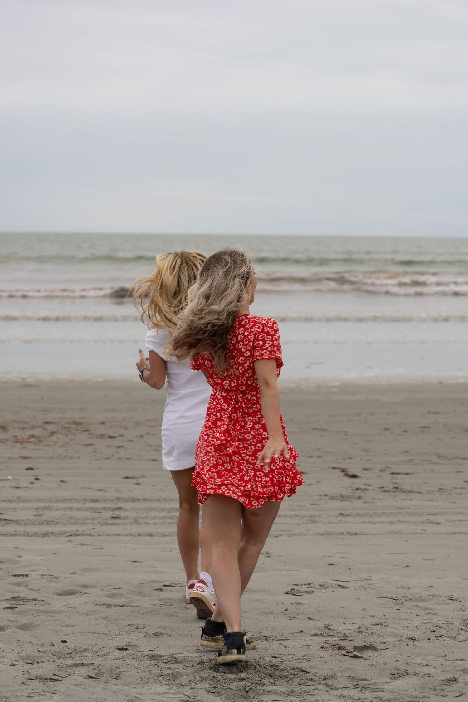 Woman in red floral dress carrying child in white on sandy beach with ocean waves in background.