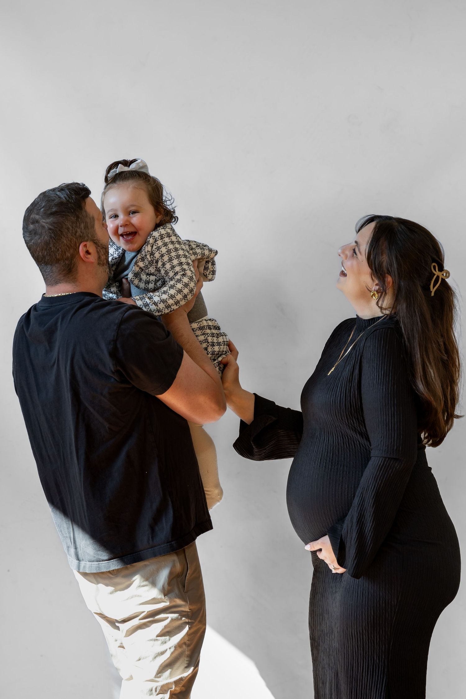 Happy family maternity photo with tattooed dad lifting laughing toddler girl while pregnant mom smiles.