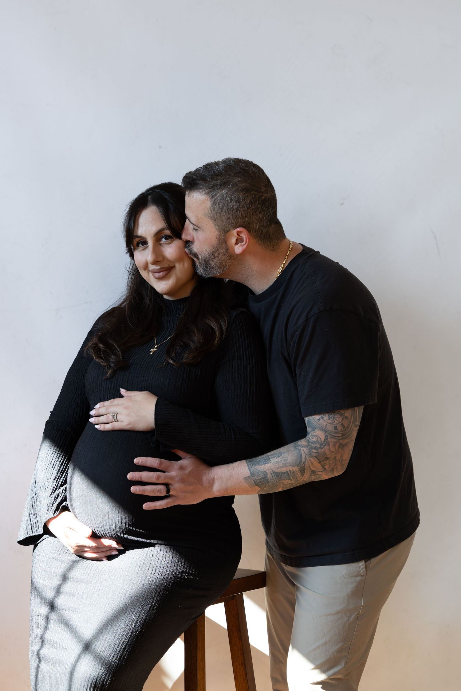 A man kisses a smiling pregnant woman on the cheek while they pose together in a bright studio setting.