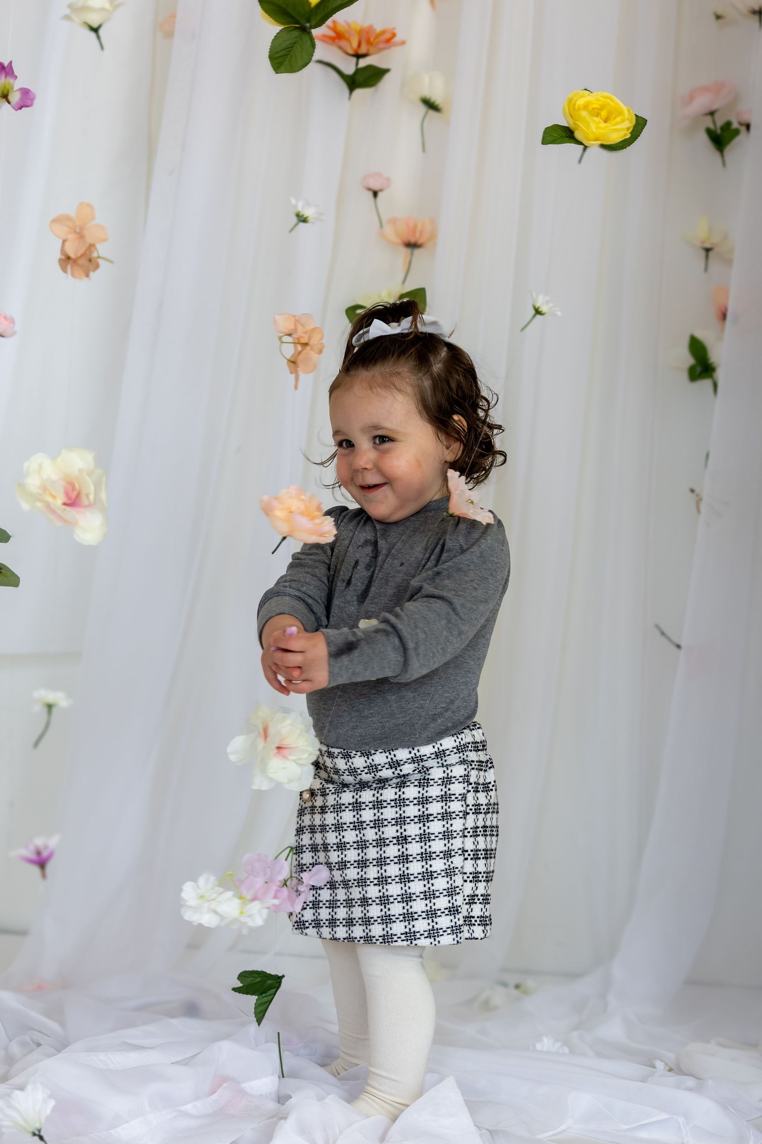 Smiling toddler girl in gray sweater and houndstooth skirt playing with flowers against white floral backdrop.