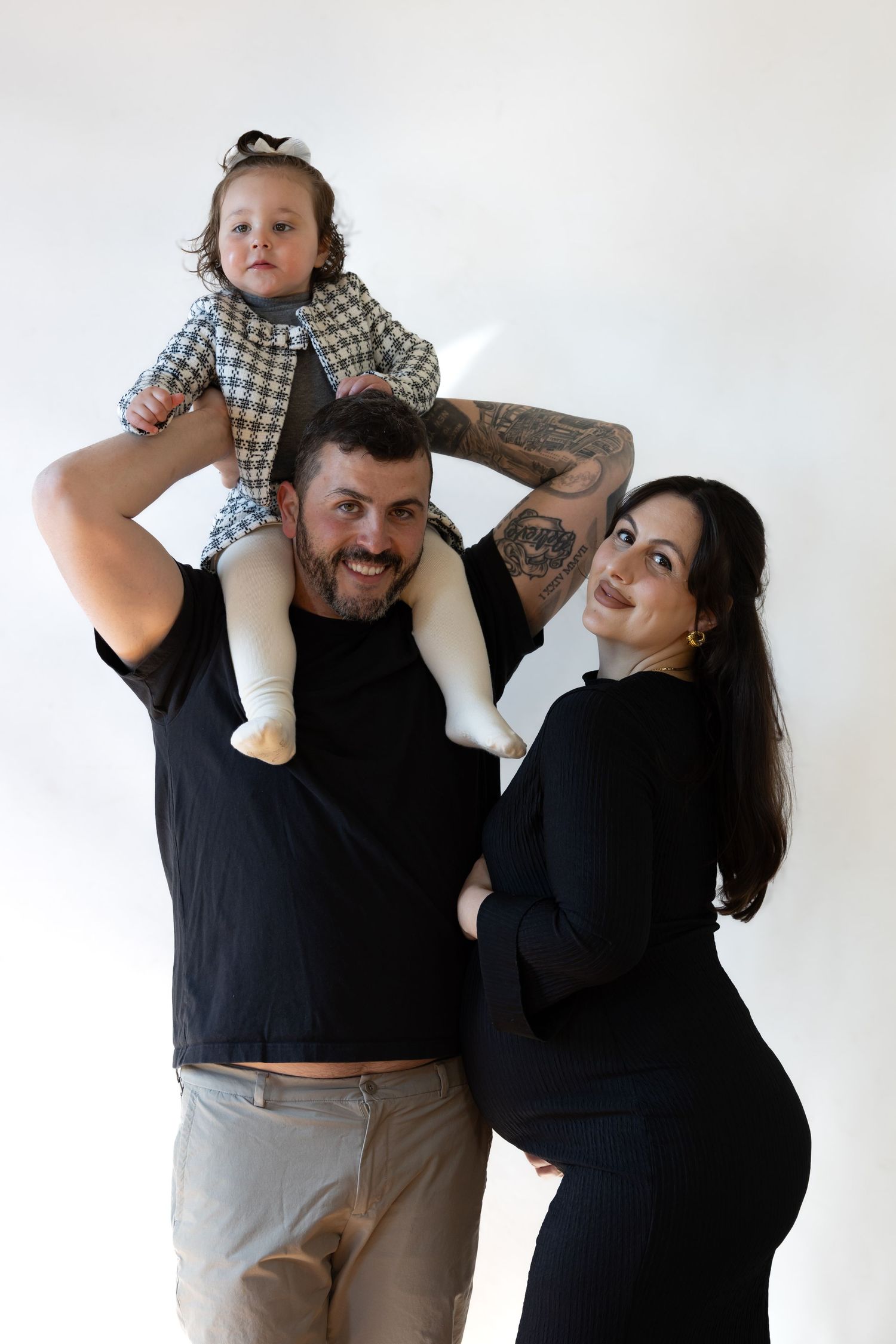 Happy family portrait with tattooed dad holding baby on shoulders beside pregnant mom in black dress.
