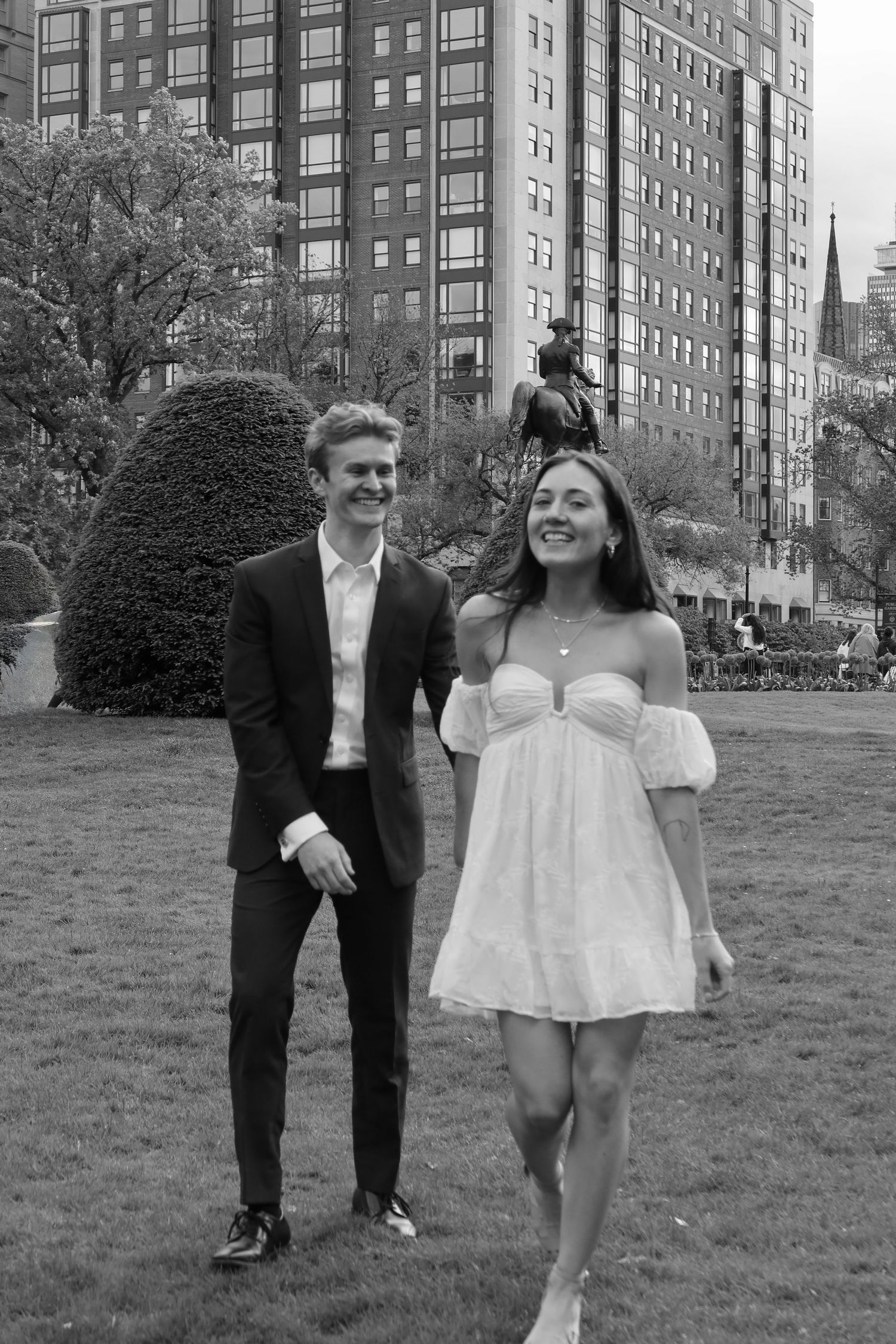 A couple in formal attire stands on a grassy lawn with tall apartment buildings in the background.