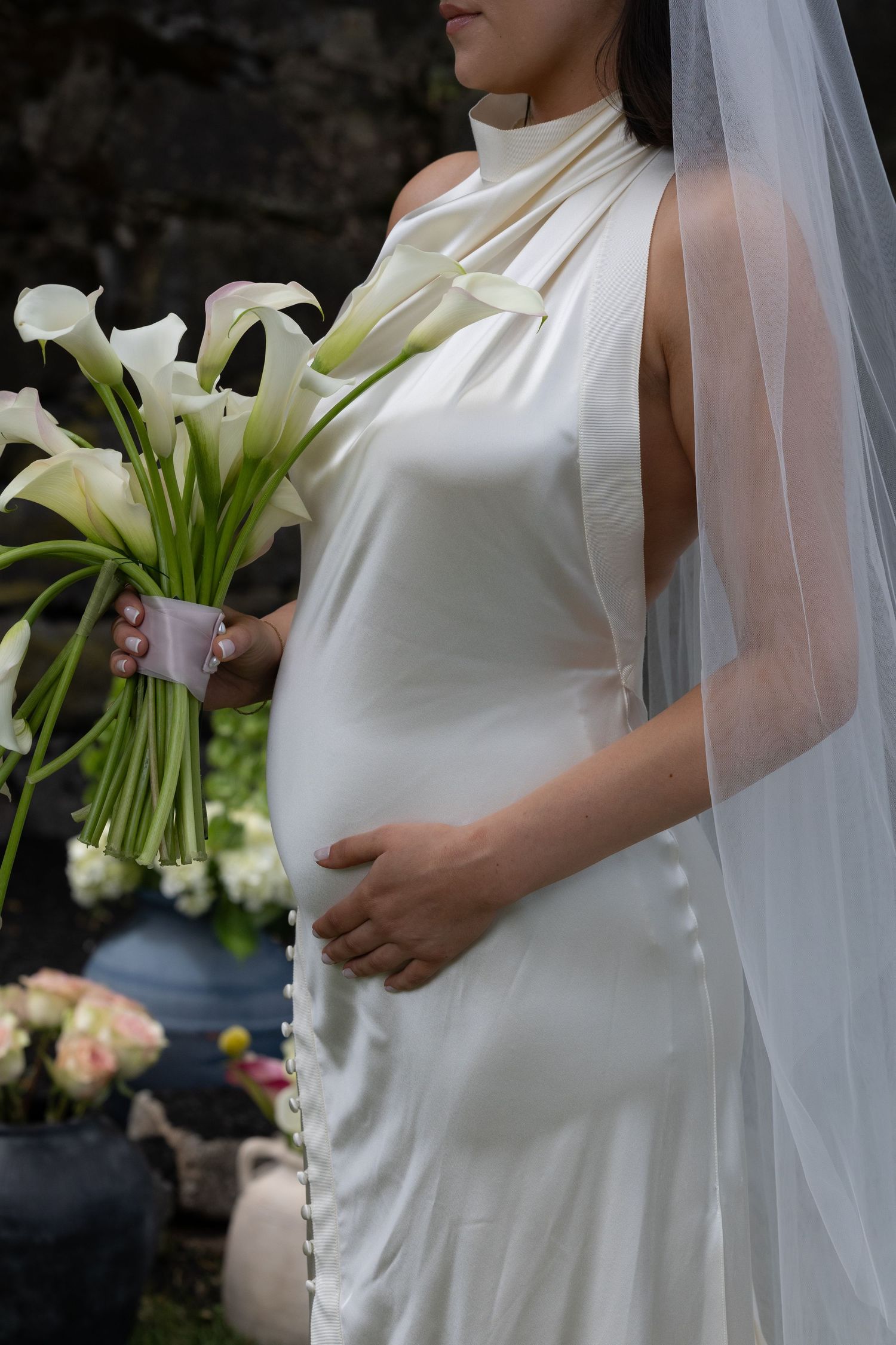 Pregnant bride in white satin halter gown holding calla lily bouquet with veil, touching baby bump.