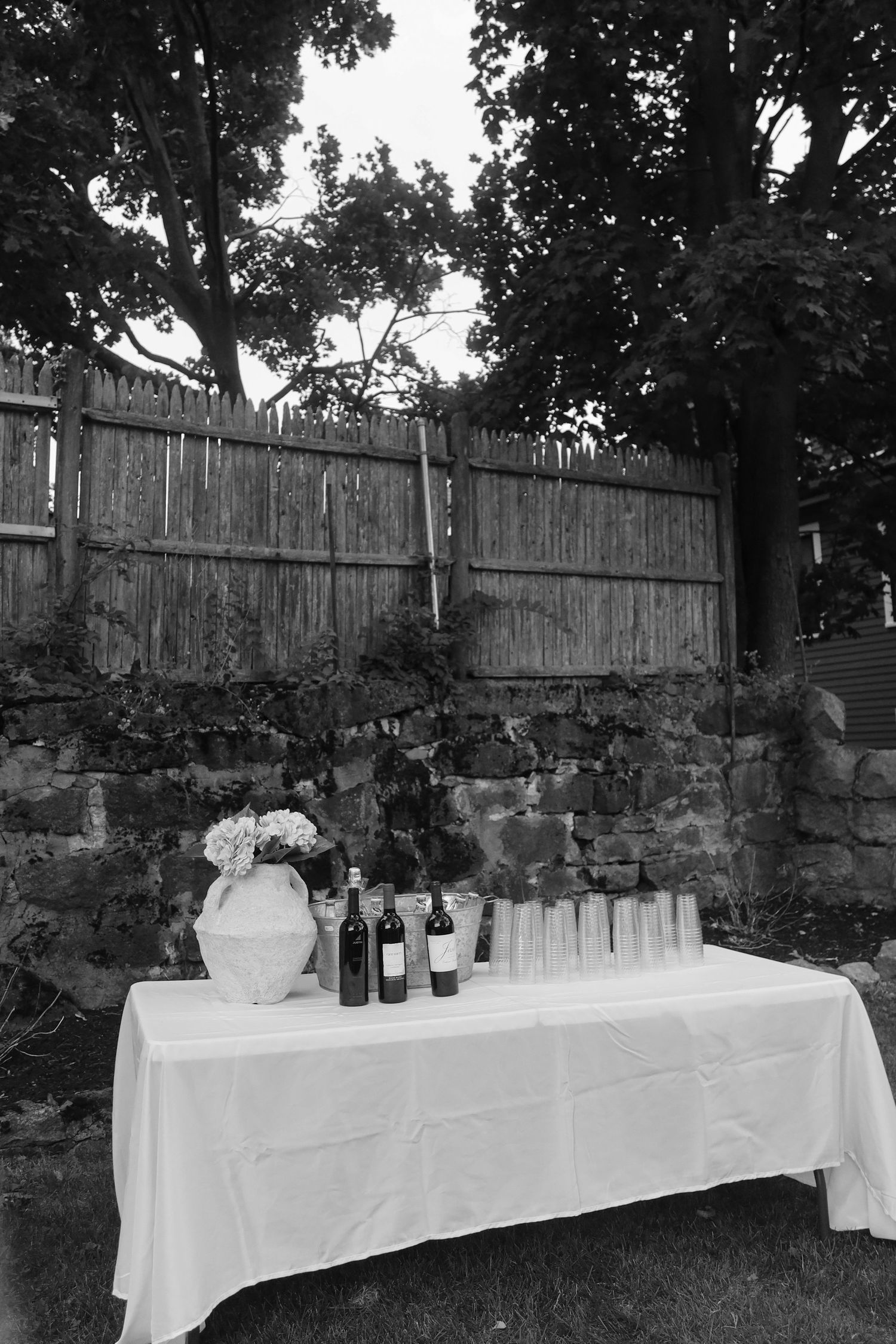 A white-clothed table with wine bottles set up against a wooden fence outdoors.