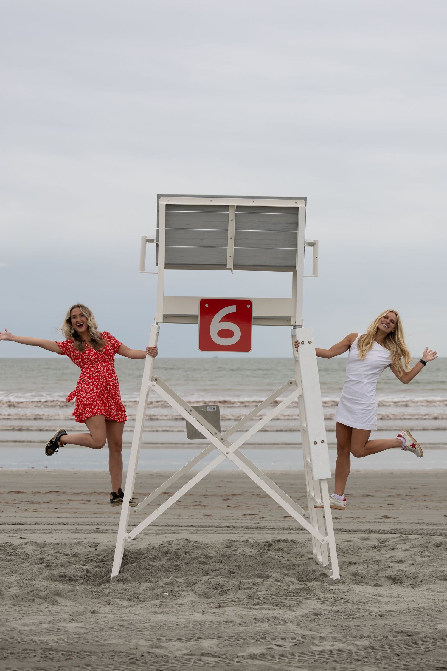 Two women in summer dresses pose joyfully beside a white lifeguard tower number 6 on a sandy beach.