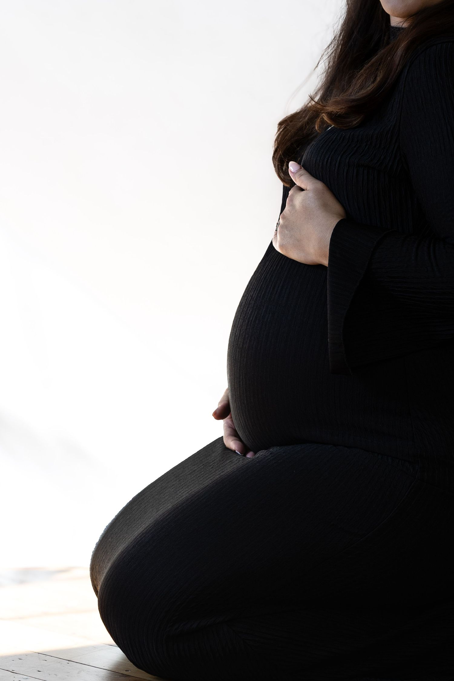 Pregnant woman in black outfit sitting cross-legged, cradling her baby bump with both hands against white background.