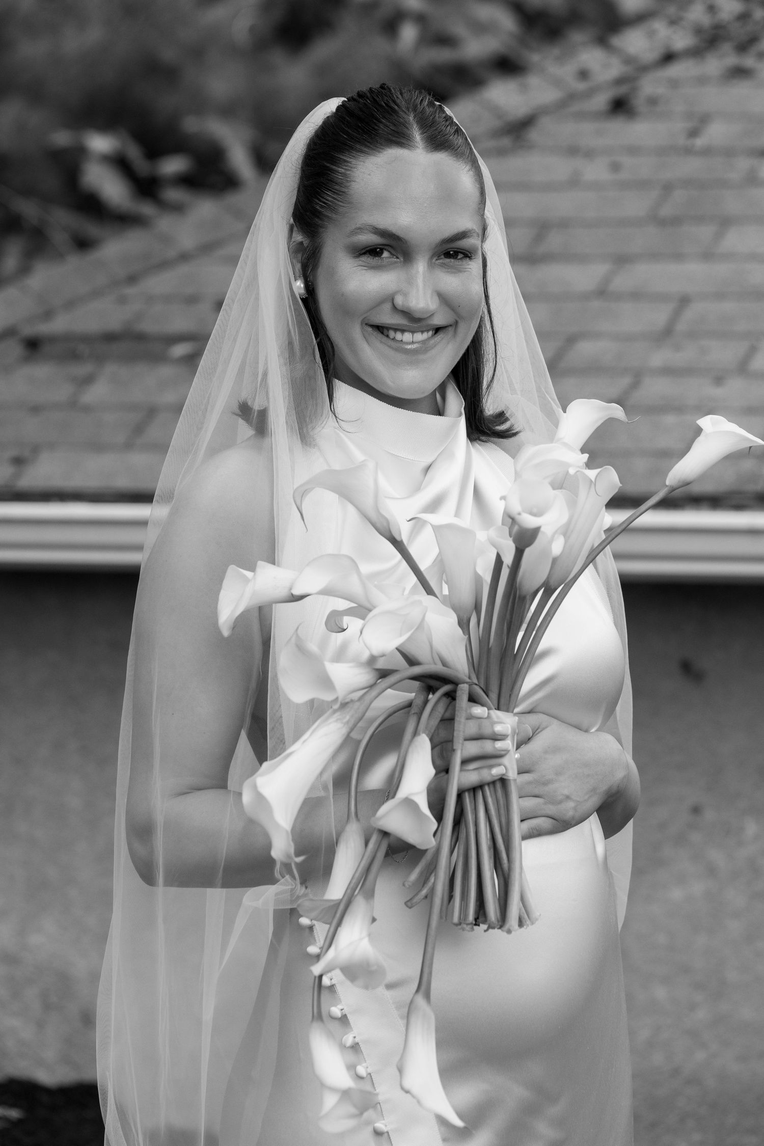 Smiling pregnant bride in white gown and veil holding calla lily bouquet, black and white wedding portrait.