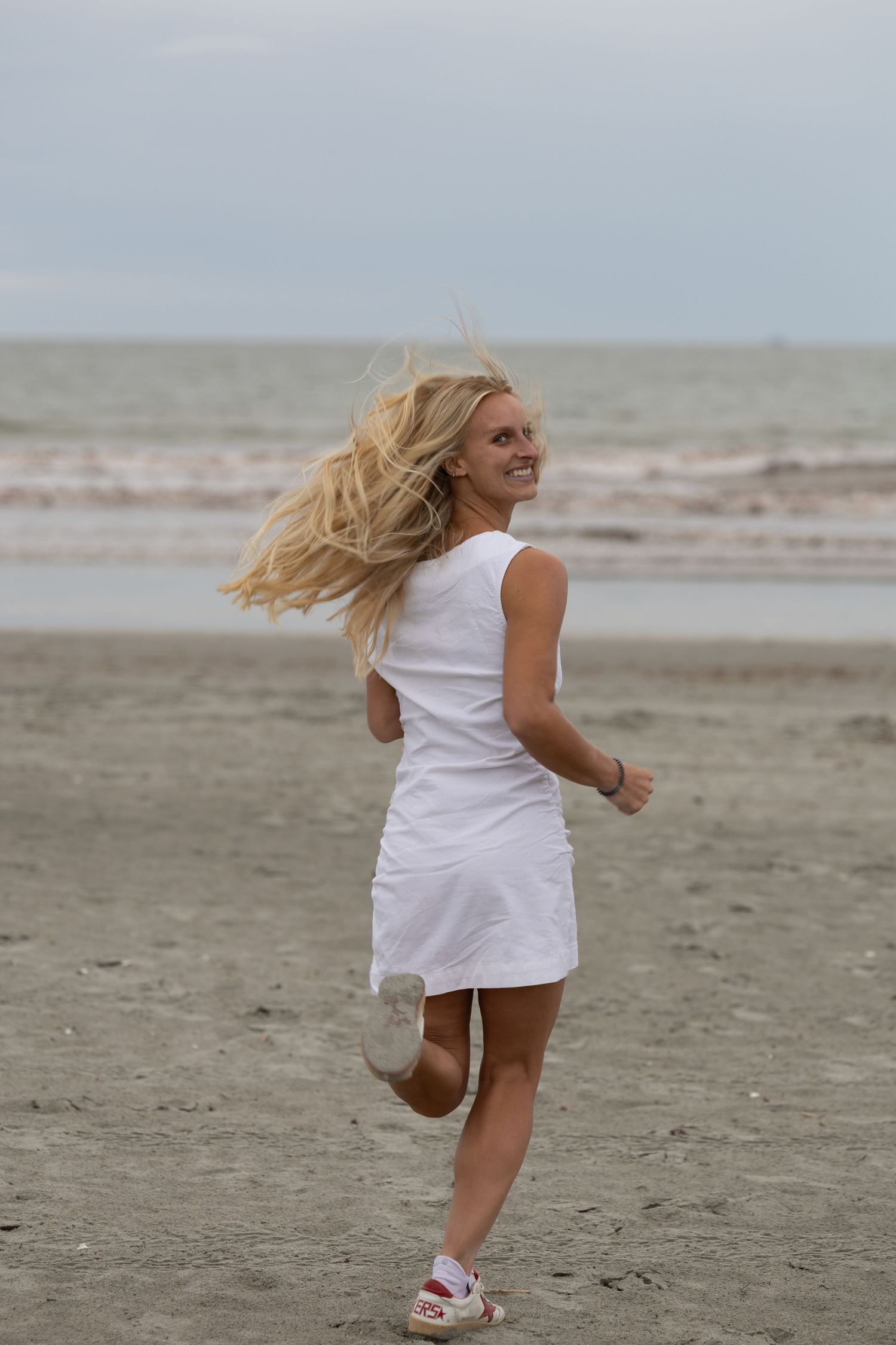 Smiling blonde woman in white dress running joyfully on a sandy beach with hair flowing in the wind.