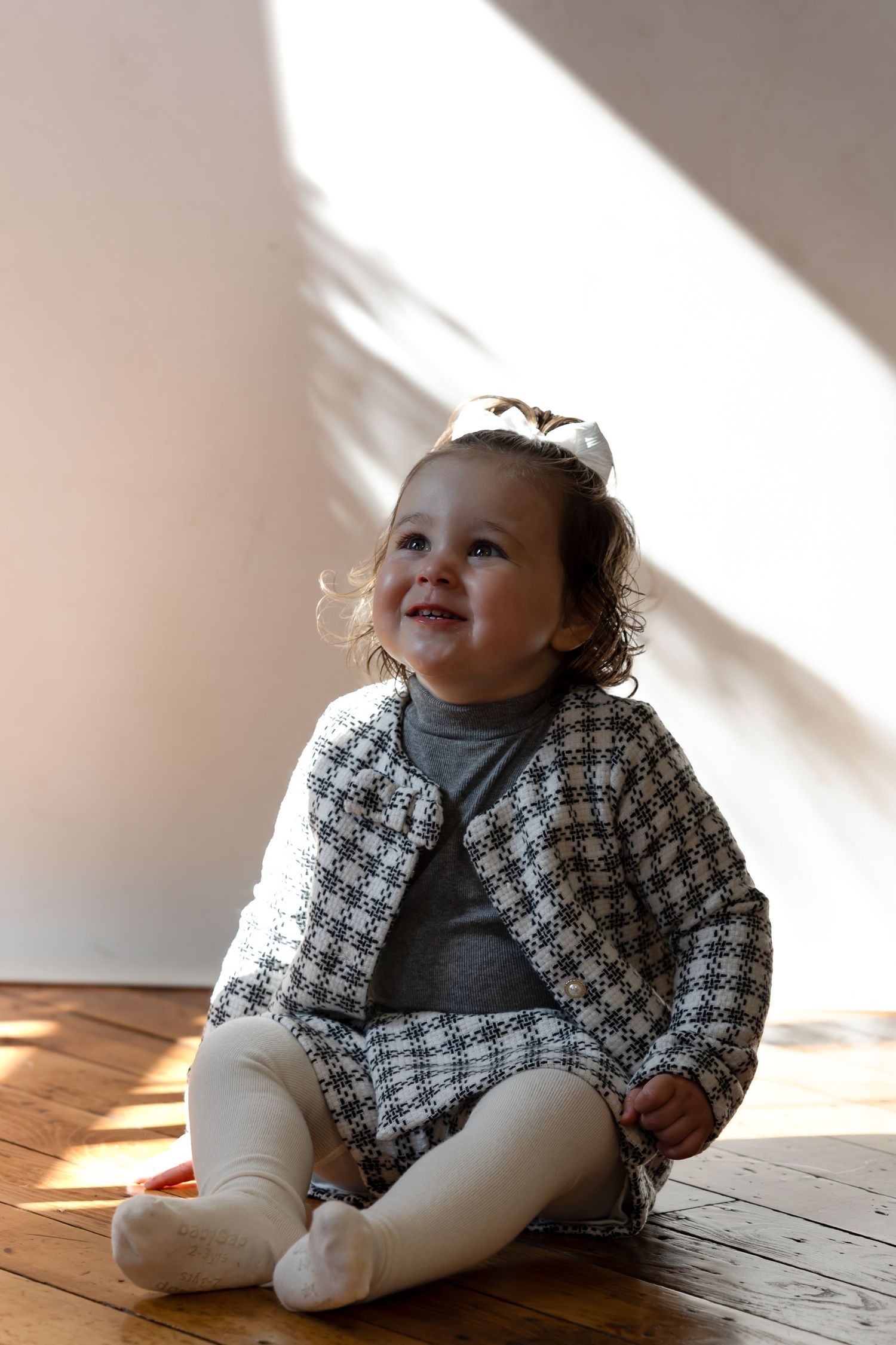 Smiling toddler girl in a plaid jacket and white tights sitting on a hardwood floor in natural light.