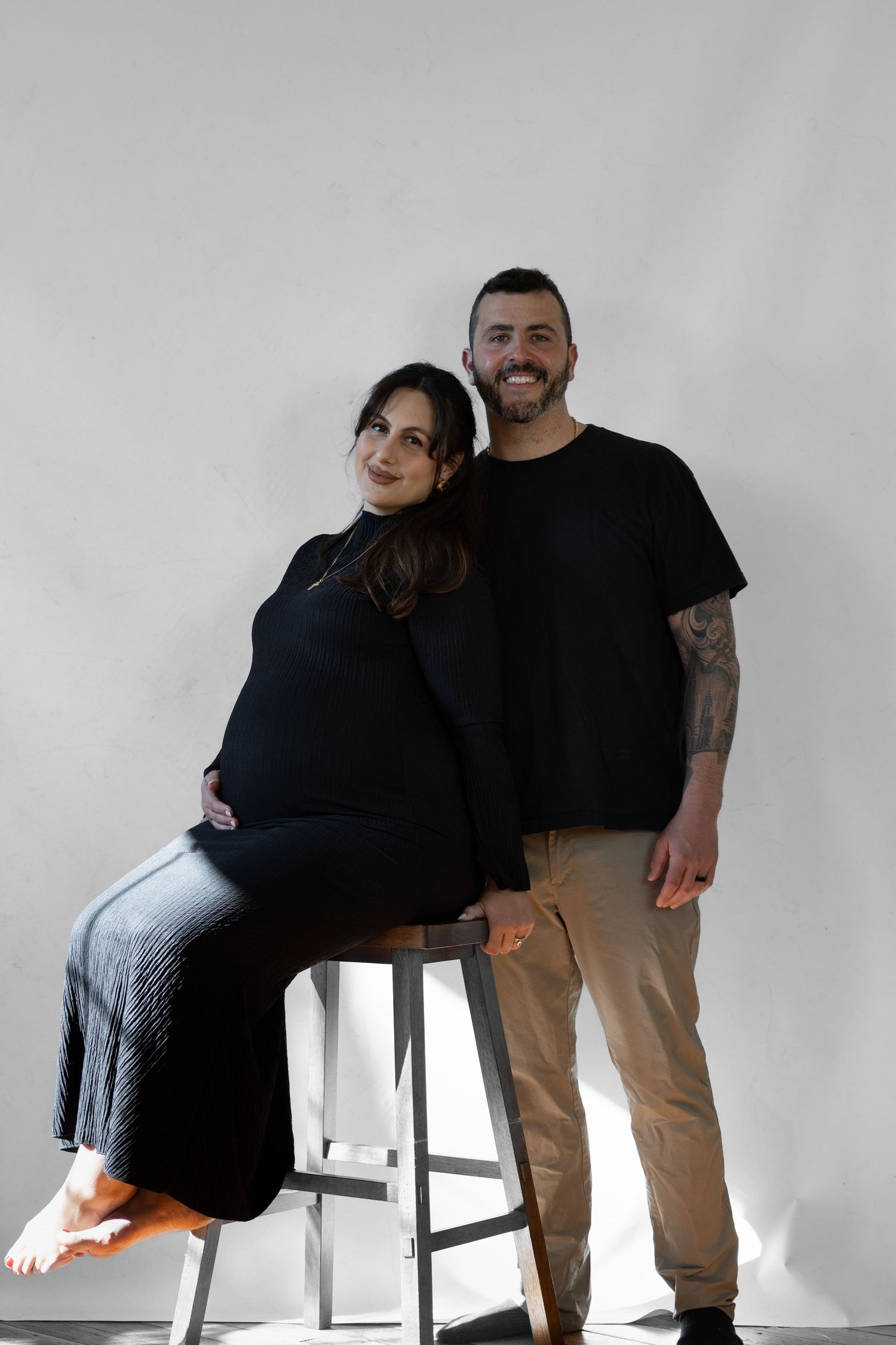 Pregnant woman in black outfit sitting on stool leaning on smiling tattooed partner in black tee against white wall.