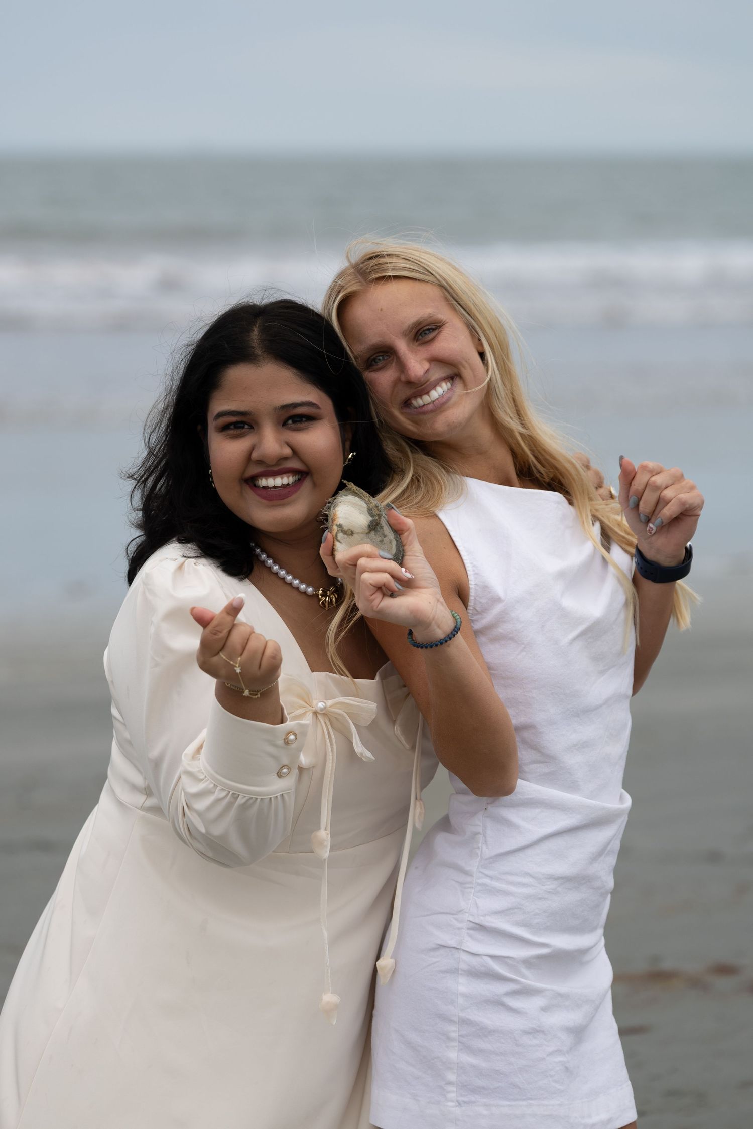 Two smiling women in white dresses pose on a beach, one holding a small shell toward the camera.